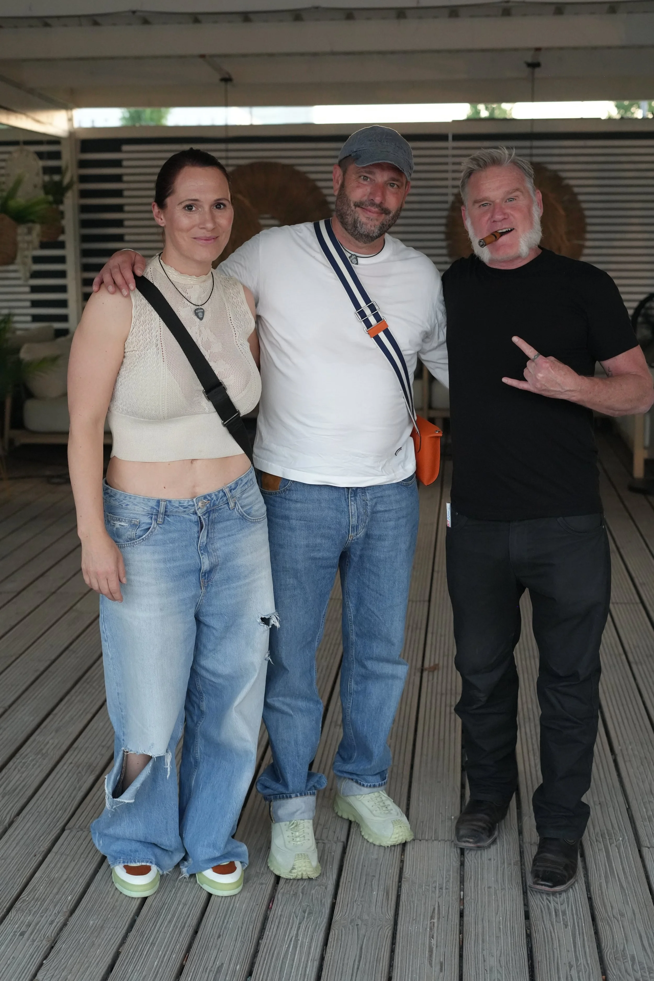 Three people standing on a wooden deck, two men and one woman, smiling at the camera. The woman is wearing a beige sleeveless crop top, ripped jeans, and colorful sneakers. The man in the middle is wearing a white T-shirt, jeans, and a gray cap with 