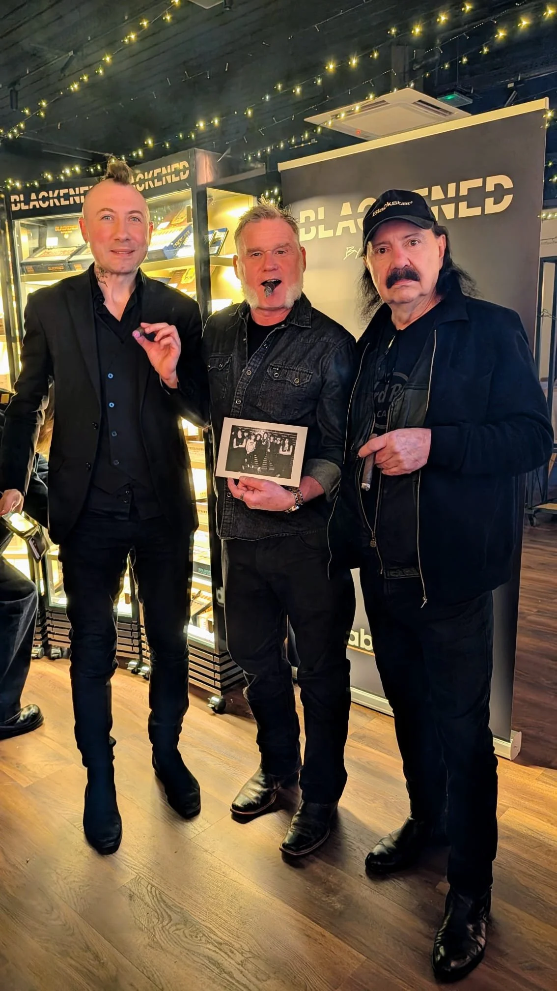 Three men standing inside a store with a sign that reads 'Blackened'. The man in the middle is holding a black and white photo. The store has string lights on the ceiling and a glass display case in the background.