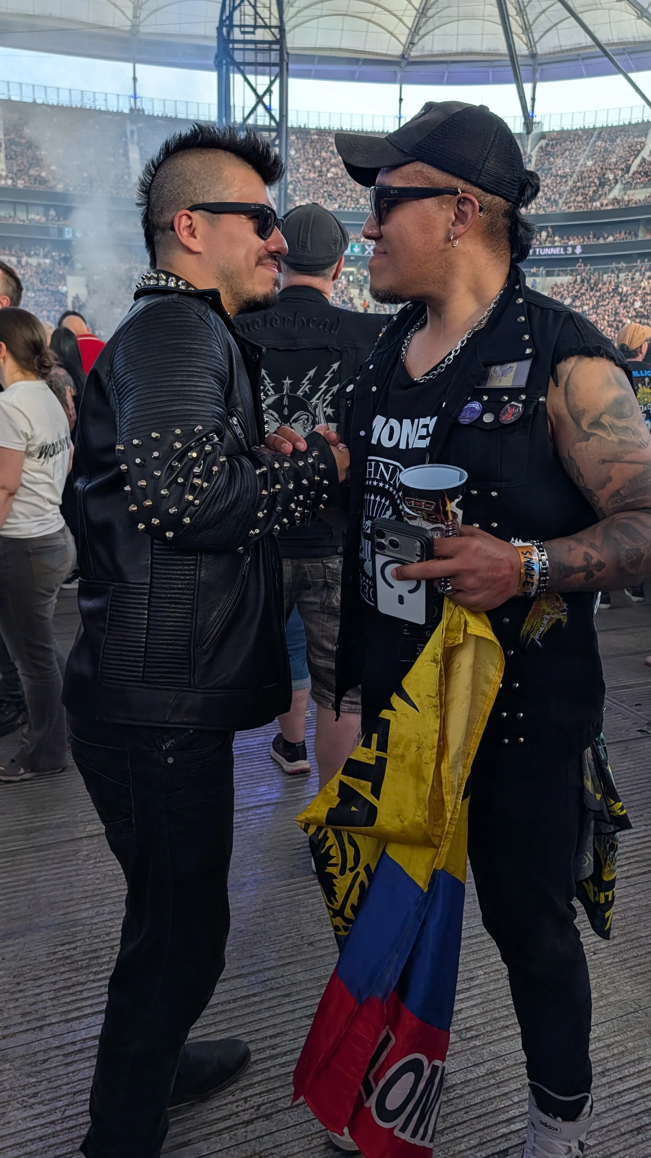 Two men facing each other and holding hands in a handshake at a concert or stadium. Both are wearing black leather jackets with spikes, sunglasses, and have tattoos. The man on the right is holding a cup and a smartphone, with a flag draped over his 