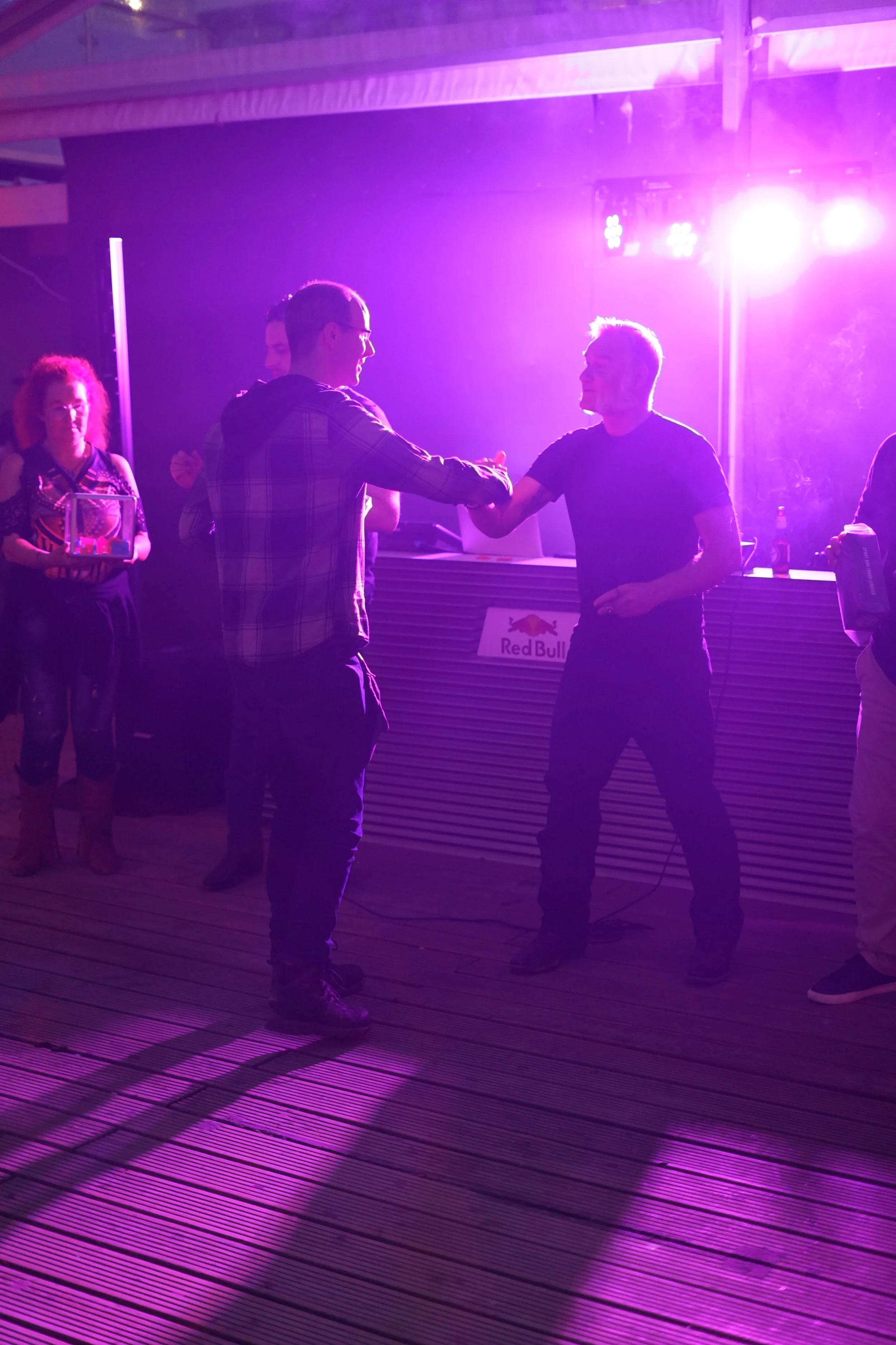 Two men shaking hands in a dimly lit room with purple lighting, with a woman holding a tray of glasses in the background and a Red Bull logo on the bar.