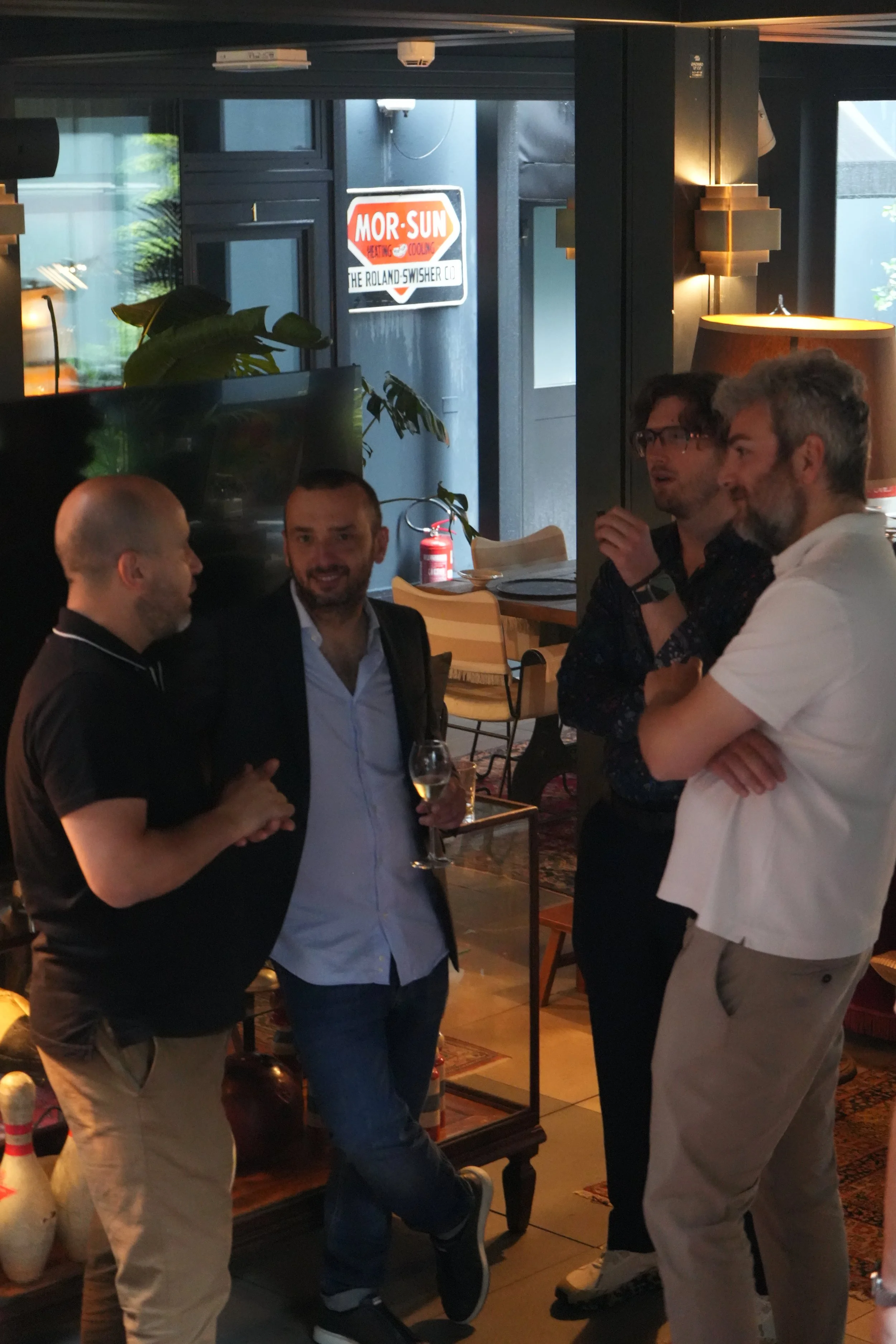 A group of four men engaged in conversation inside a restaurant or cafe, with a sign outside the window reading 'MOR-SUN Heating & Cooling The Roland Swisher Co.' and decorative plants visible in the background.