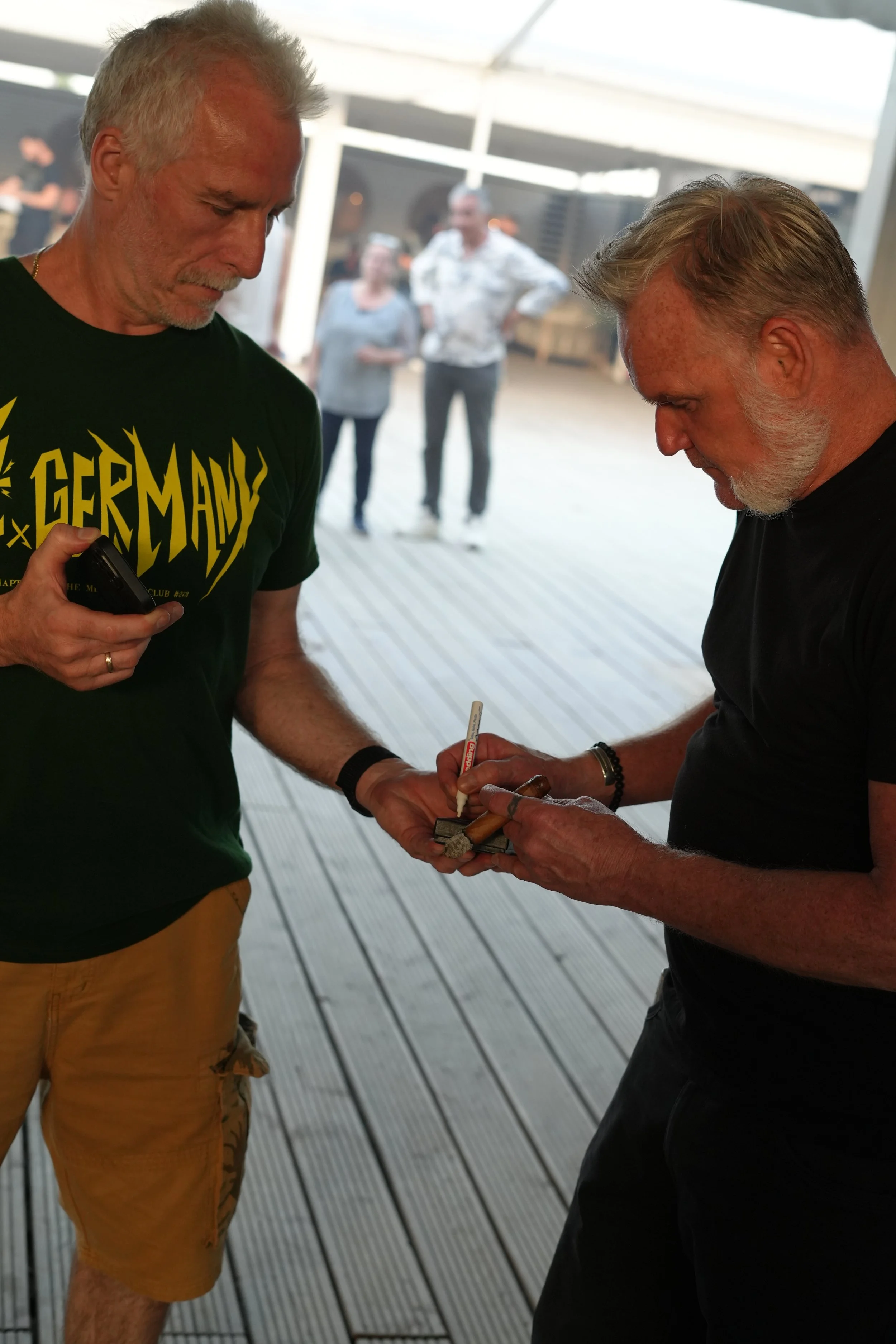 Two men with gray hair and beards signing a small wooden object in an outdoor covered area, with two women observing in the background.
