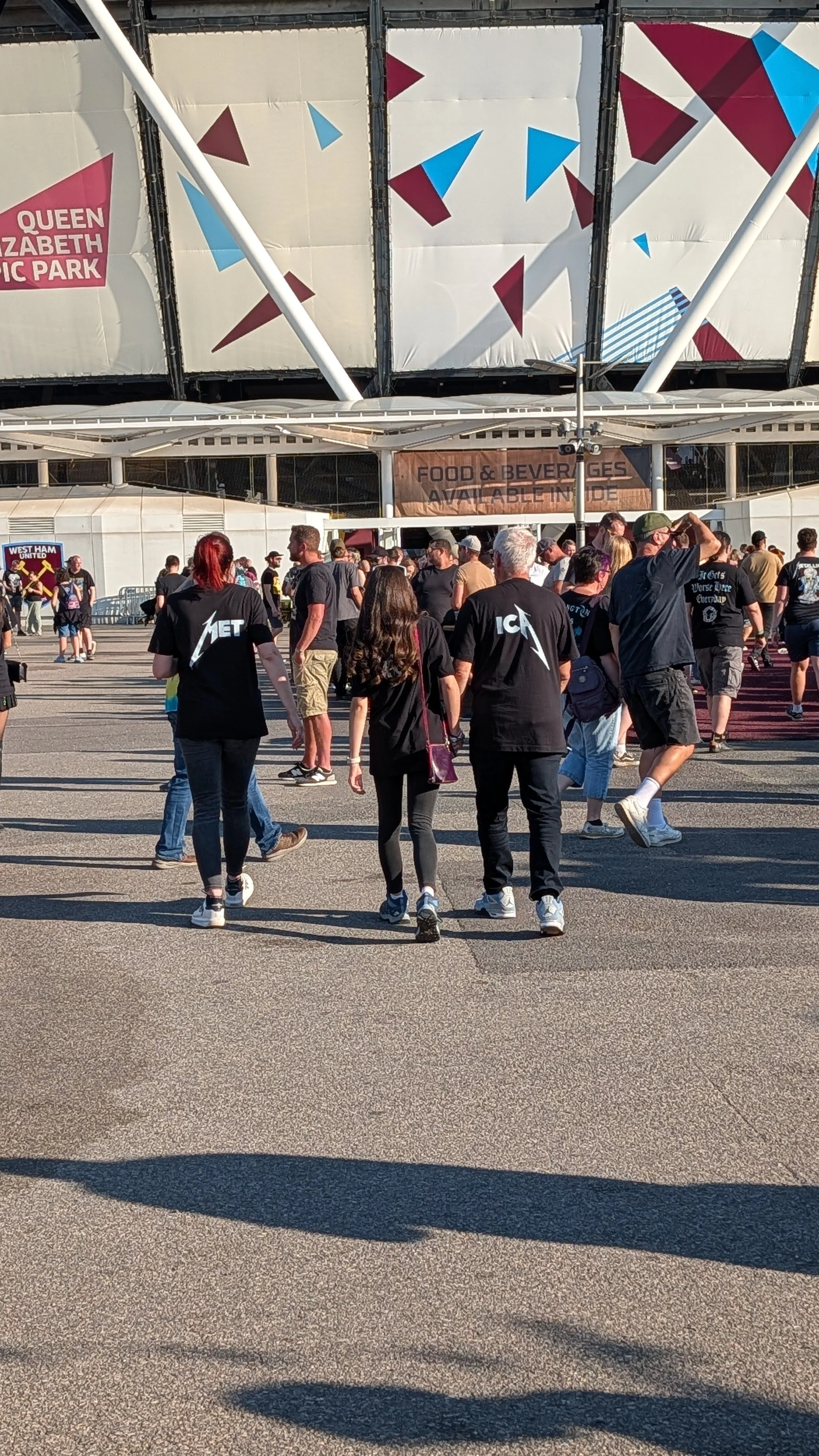 Crowd of people walking outside Queen Elizabeth Olympic Park, with a large advertising banner and a building in the background.