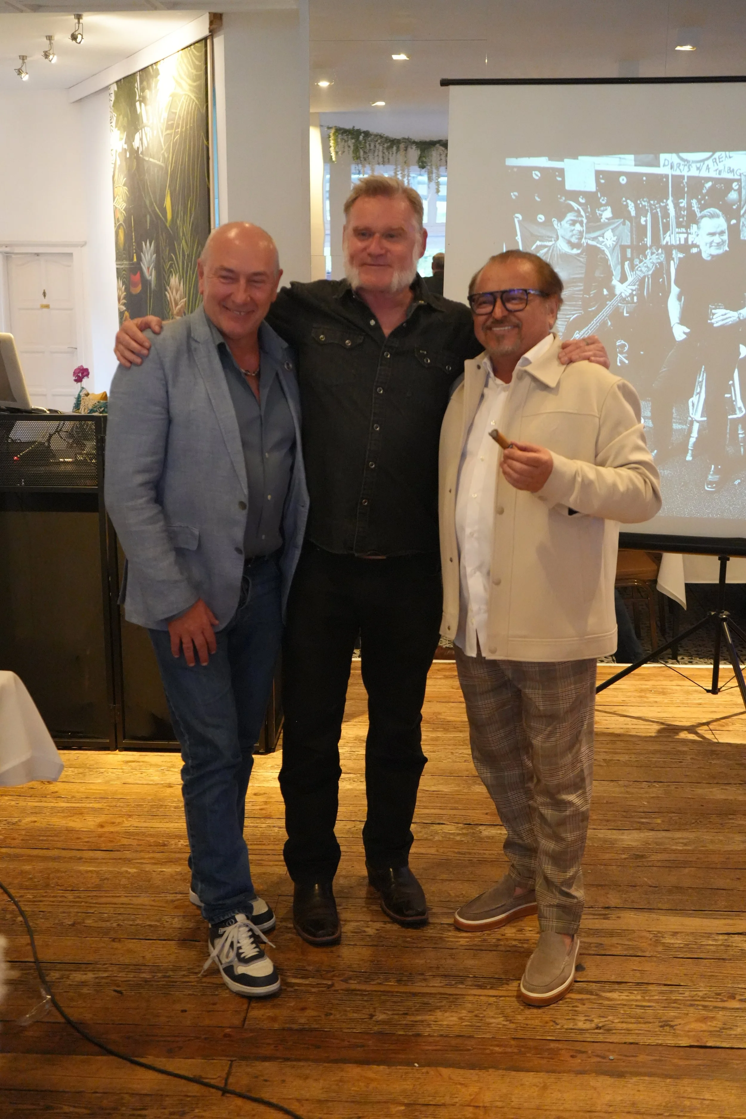 Three men standing together indoors, smiling, with a black-and-white photo projected behind them and a wooden floor underneath.