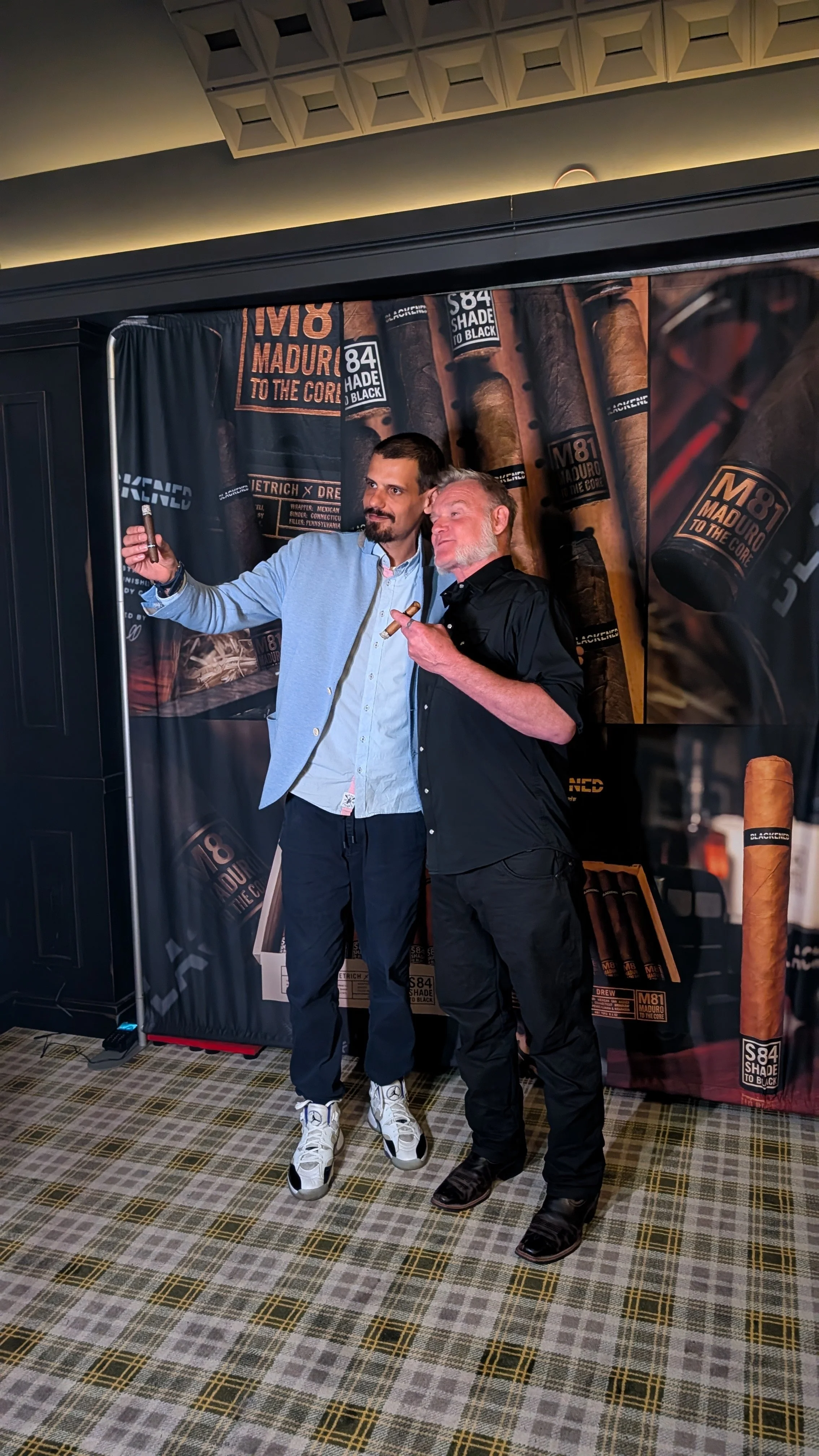 Two men taking a selfie in front of a background with cigar images. One man is wearing a light blue blazer and white sneakers, and the other is in black clothing with a cigar in hand.