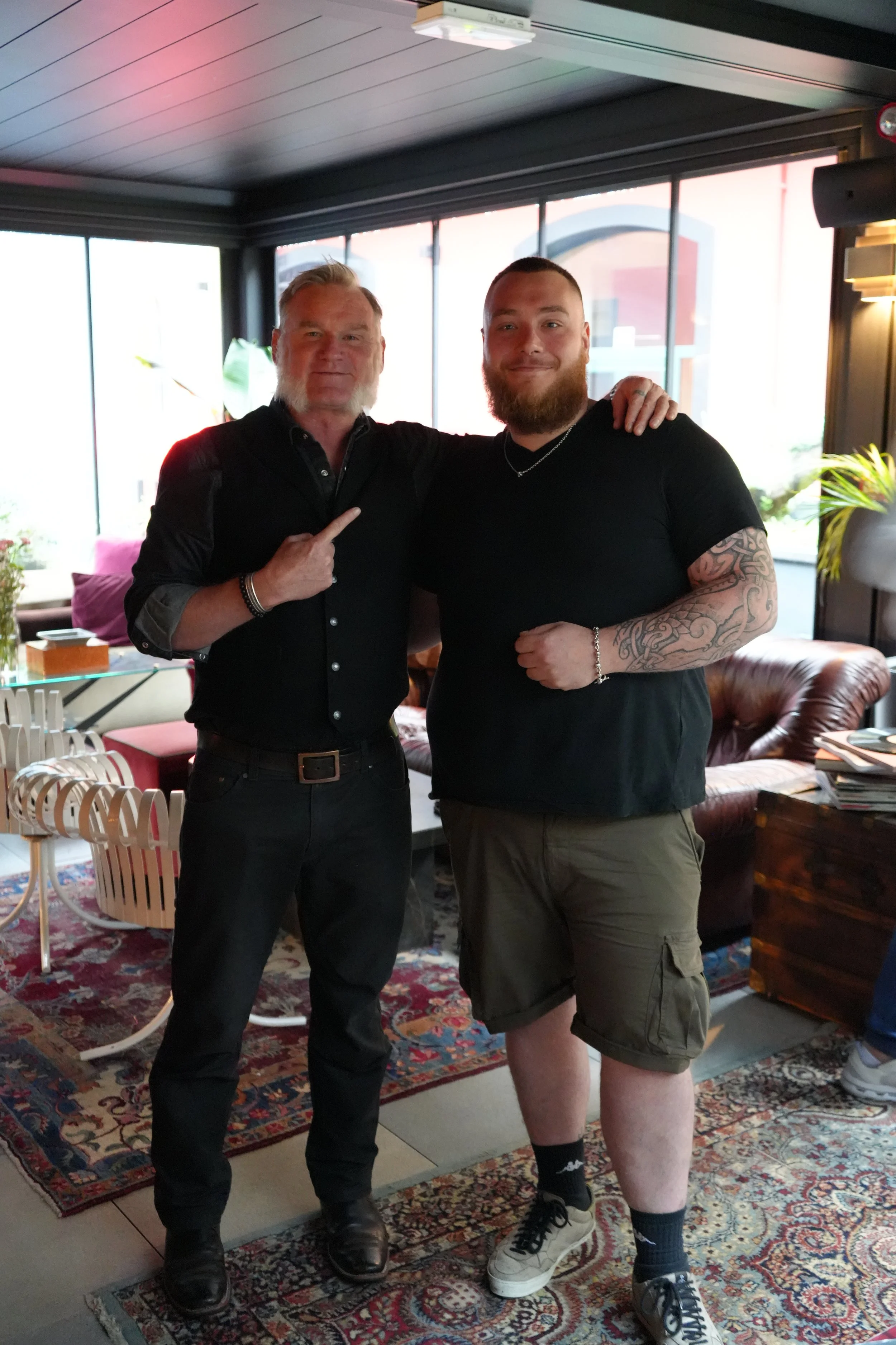 Two men standing together indoors, one with silver hair and beard pointing at the other, who has a full beard and tattoos on his arm. They are smiling. The background shows large windows, plants, a leather chair, and a patterned carpet.
