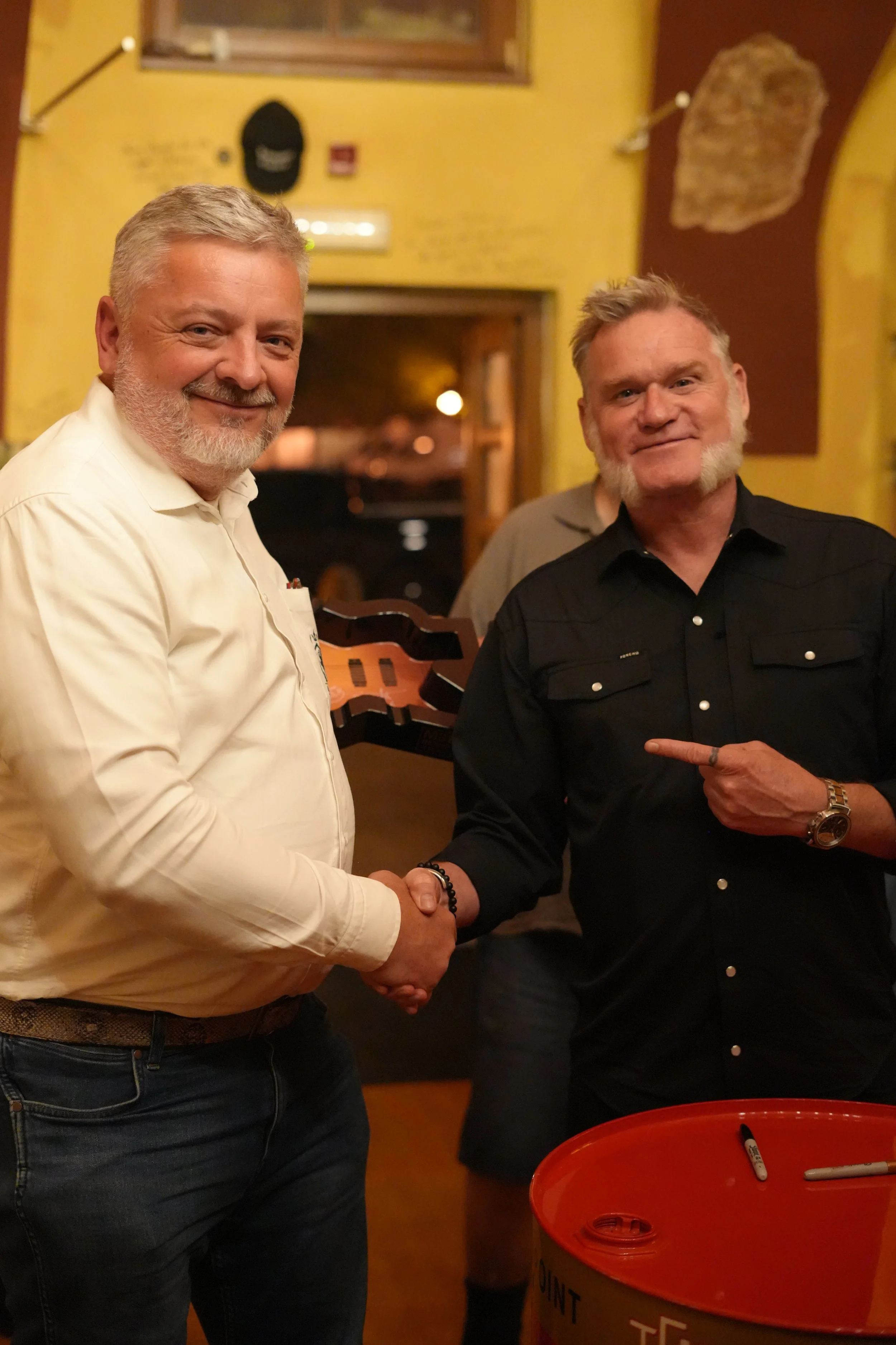 Two men shaking hands in a restaurant, with one wearing a white shirt and the other wearing a black shirt, and a red donation bin with markers on a table in the foreground.