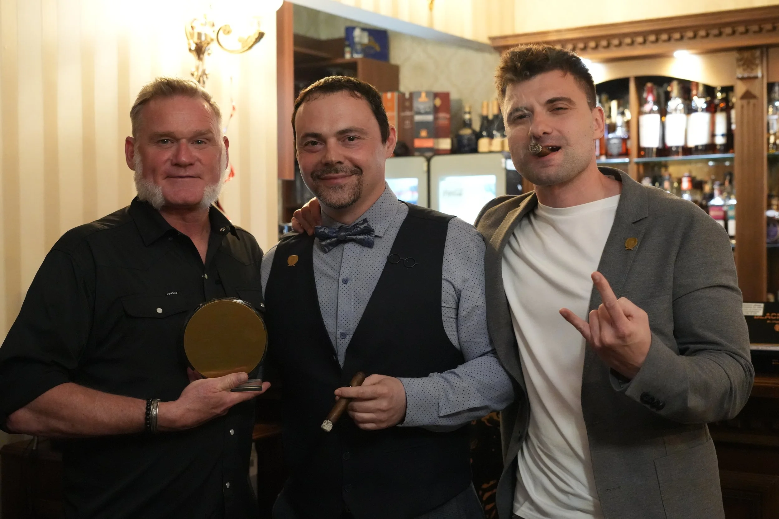 Three men standing together in a bar, celebrating. The man on the left is holding a gold-colored award or plaque. The man in the middle is smiling and wearing a patterned shirt with a bow tie. The man on the right is making a hand gesture with his fi