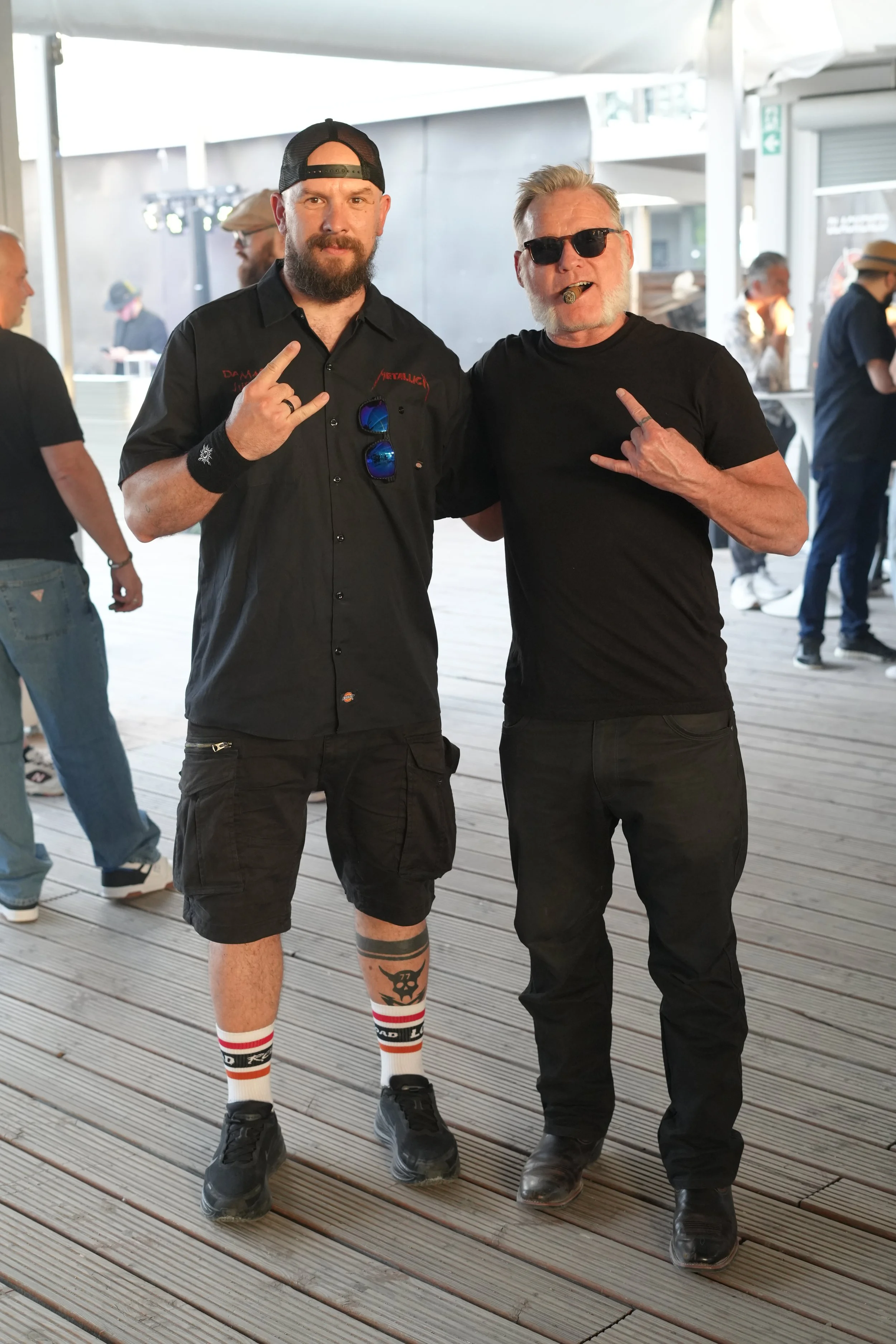 Two men posing at an outdoor event, making rock signs with their hands. One man has a beard, tattoos, a black cap, black shirt with 'Metallica' on it, and sports sunglasses. The other man has a beard, sunglasses, and a cigar in his mouth, wearing a b
