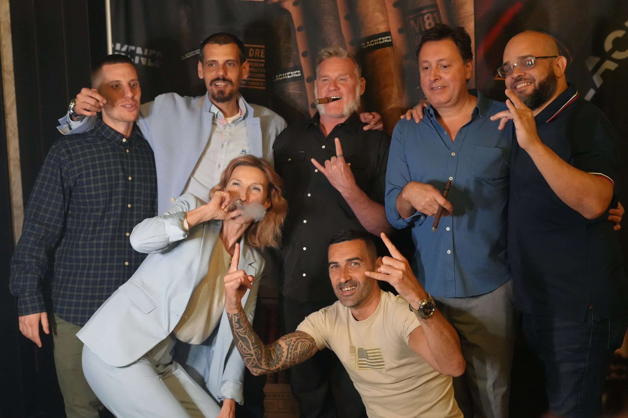 Group of six people in a social setting posing together, with some making hand gestures, smoking cigars, and smiling.