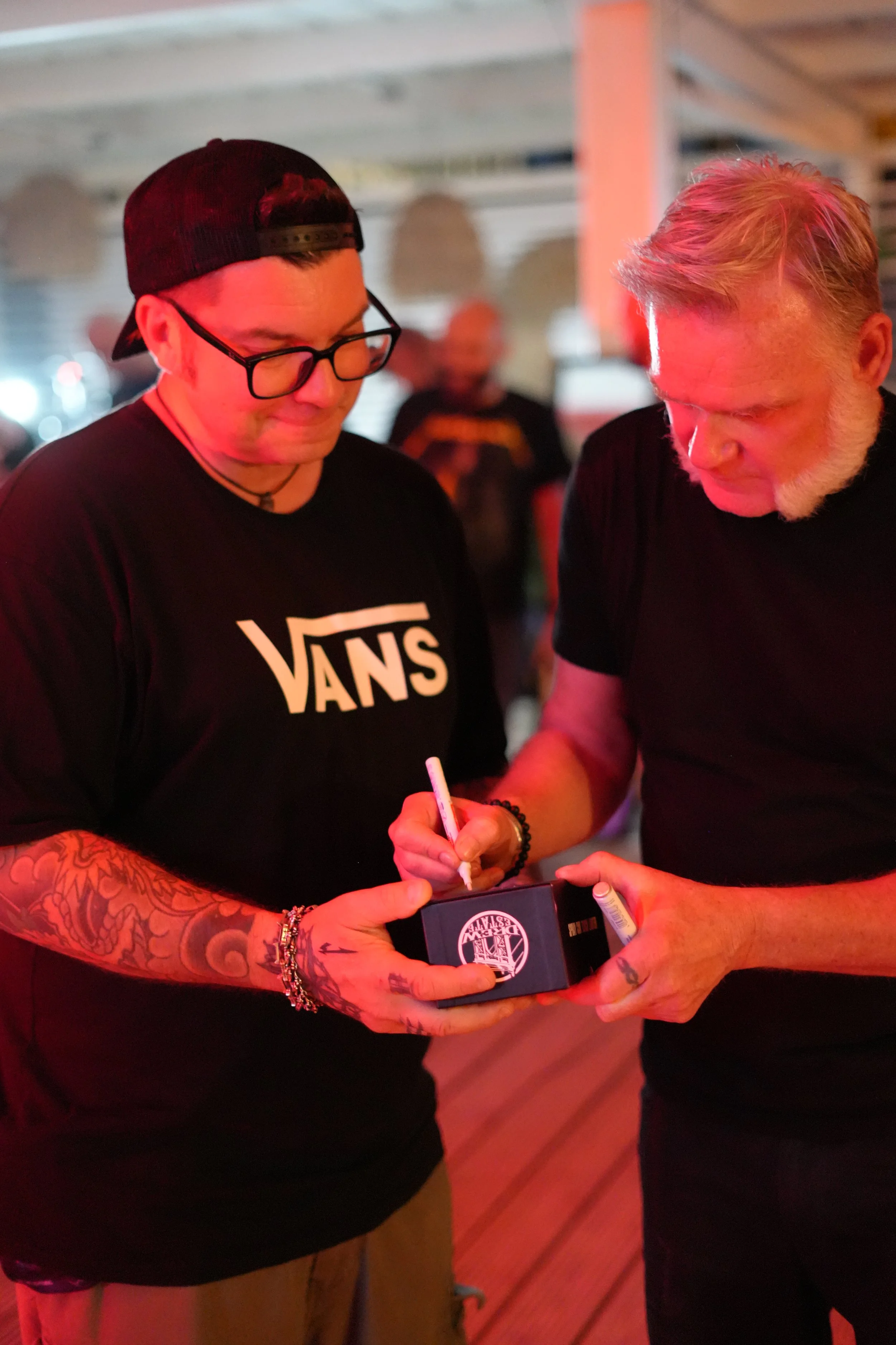 Two men with tattoos and facial hair standing close, looking at a small black box with a white logo. One man is wearing glasses, a black cap backwards, a black t-shirt with a white logo, and jewelry. The other man has blond hair and a beard, wearing 