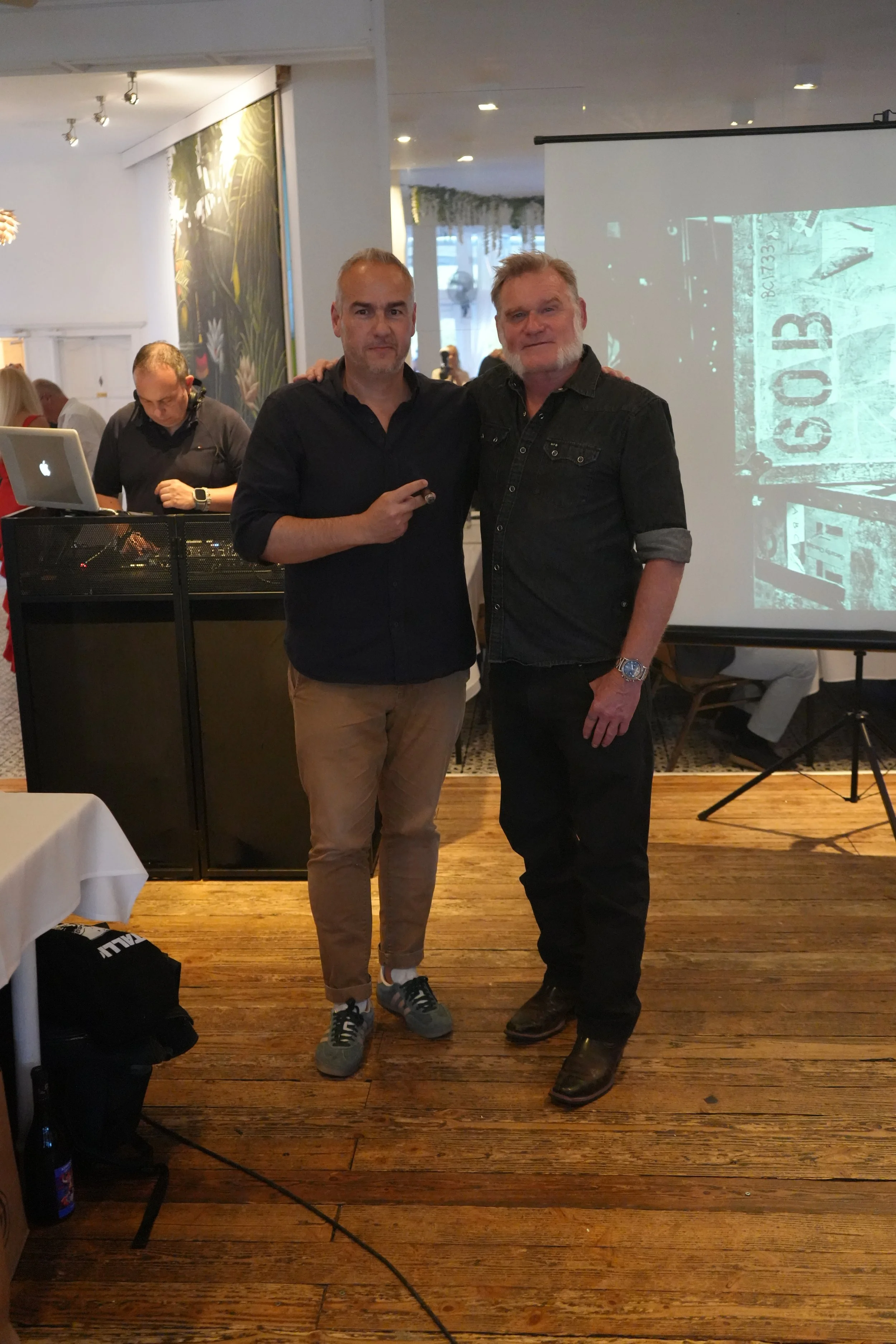 Two men standing next to each other, smiling, at an indoor event, with one giving a peace sign. A DJ is in the background, and a projection screen displays an image of a street scene with a sign that reads 'G04'.