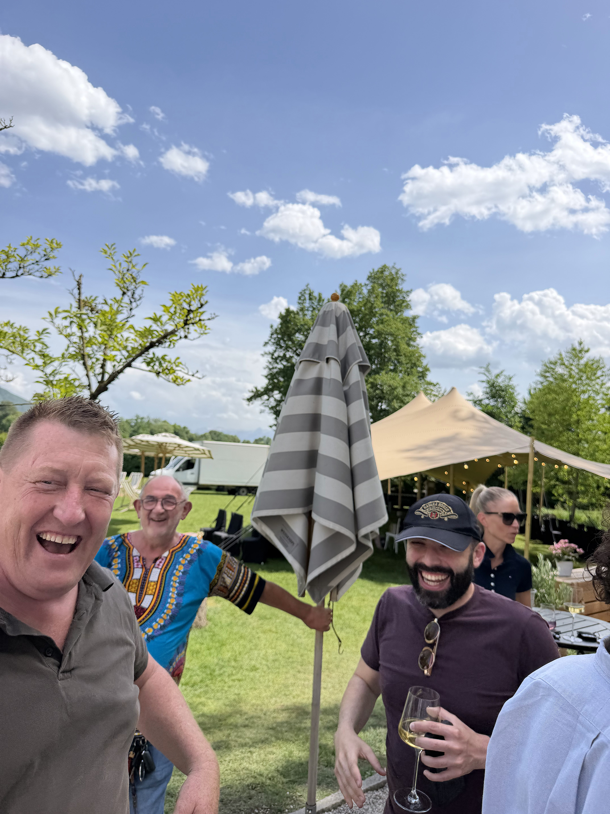 Group of friends celebrating outdoors at a backyard gathering with trees, umbrellas, and a partly cloudy sky.