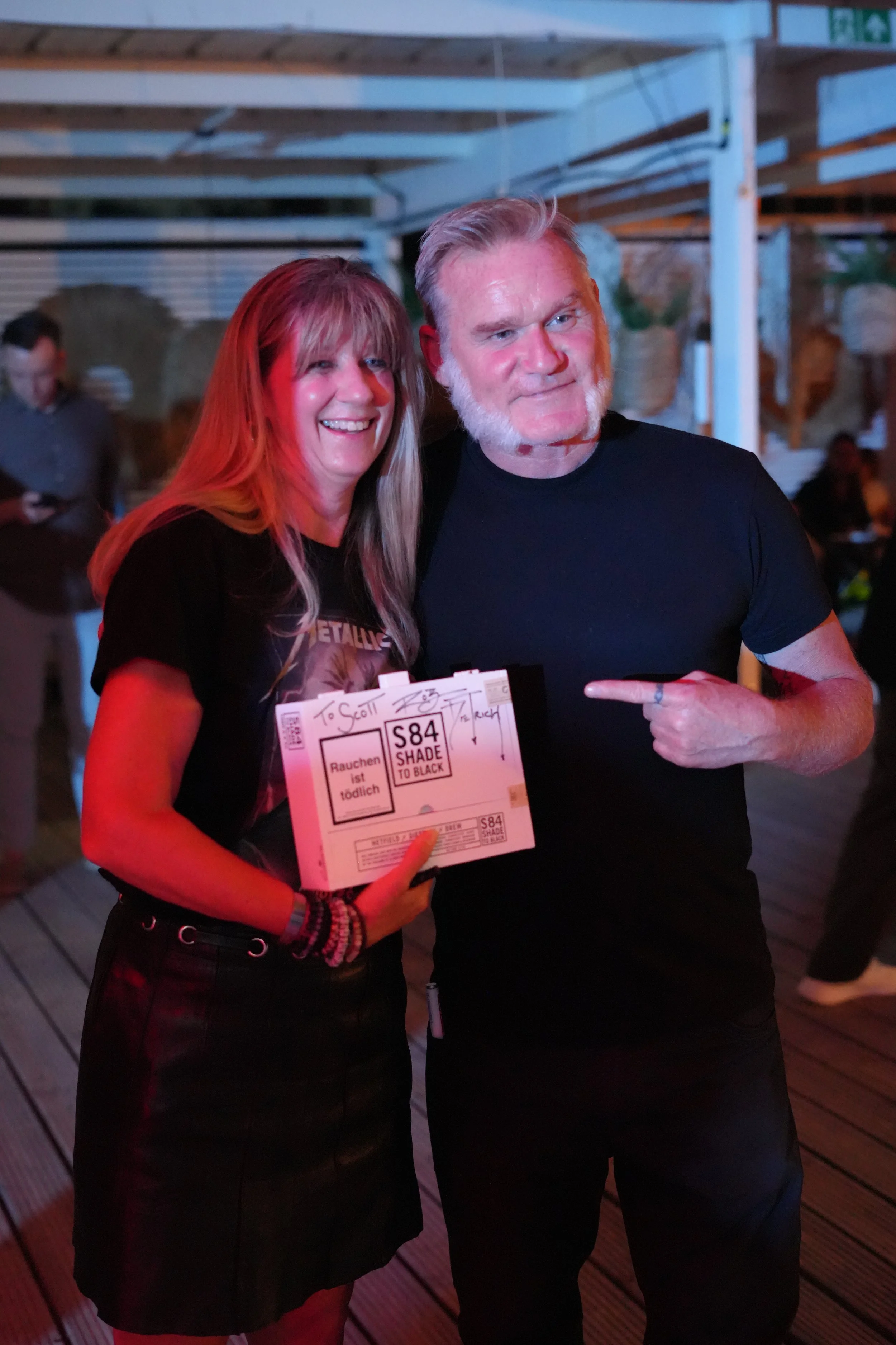 A woman and a man smiling at a social event. The woman is holding a ticket, and the man is pointing at her. The background shows other people and wooden flooring.