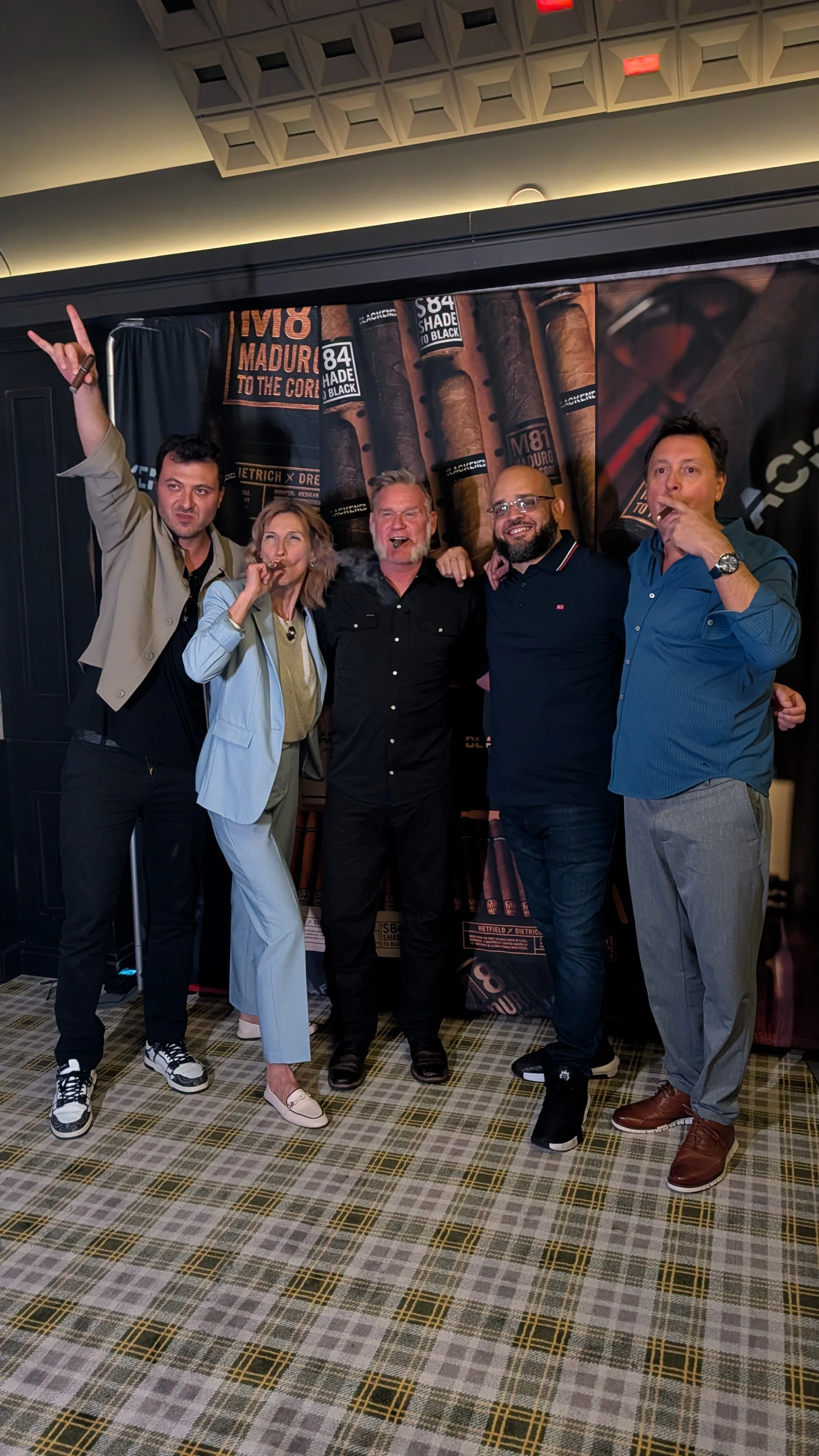 Group of five people posing at an indoor cigar event, standing in front of a large banner displaying cigars.