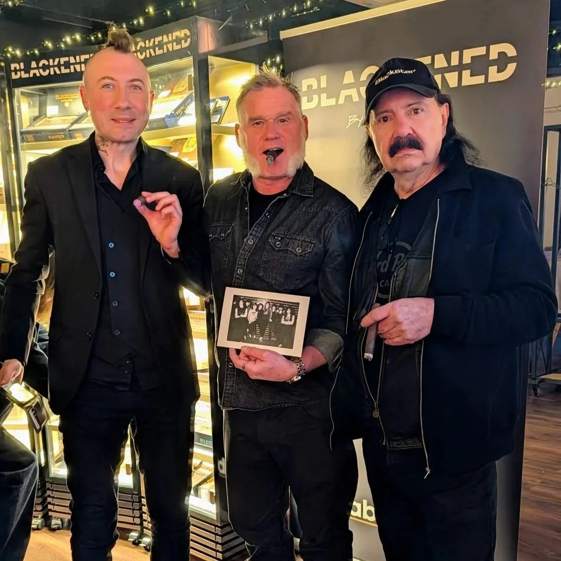 Three men posing together at an event, with one holding a photograph and another cigar, in front of a display promoting Blackened by Rob Dietrich.
