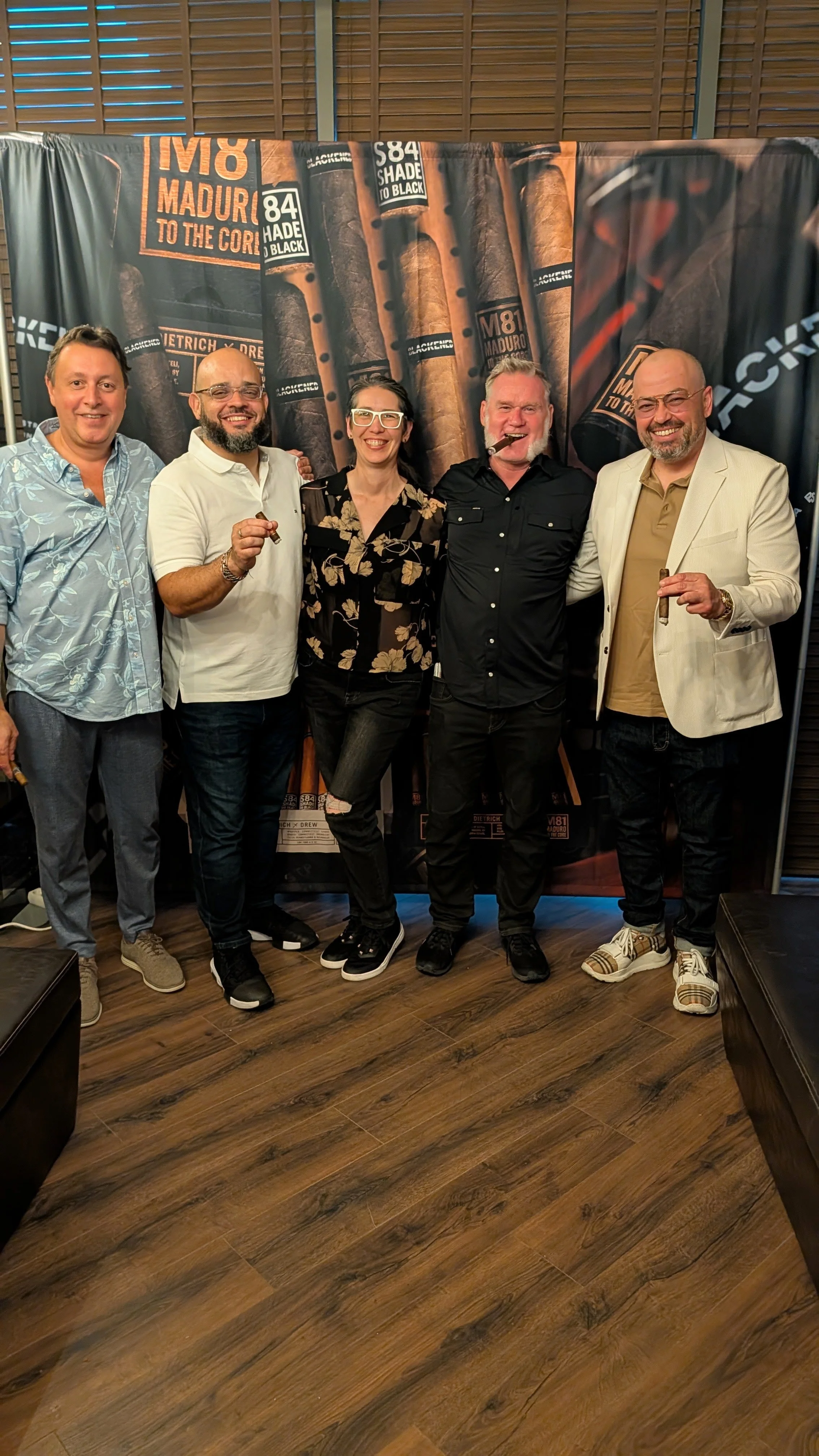 Five people standing together in a room with wood flooring and a backdrop featuring cigars and cigar labels. They are smiling, and some are holding cigars.