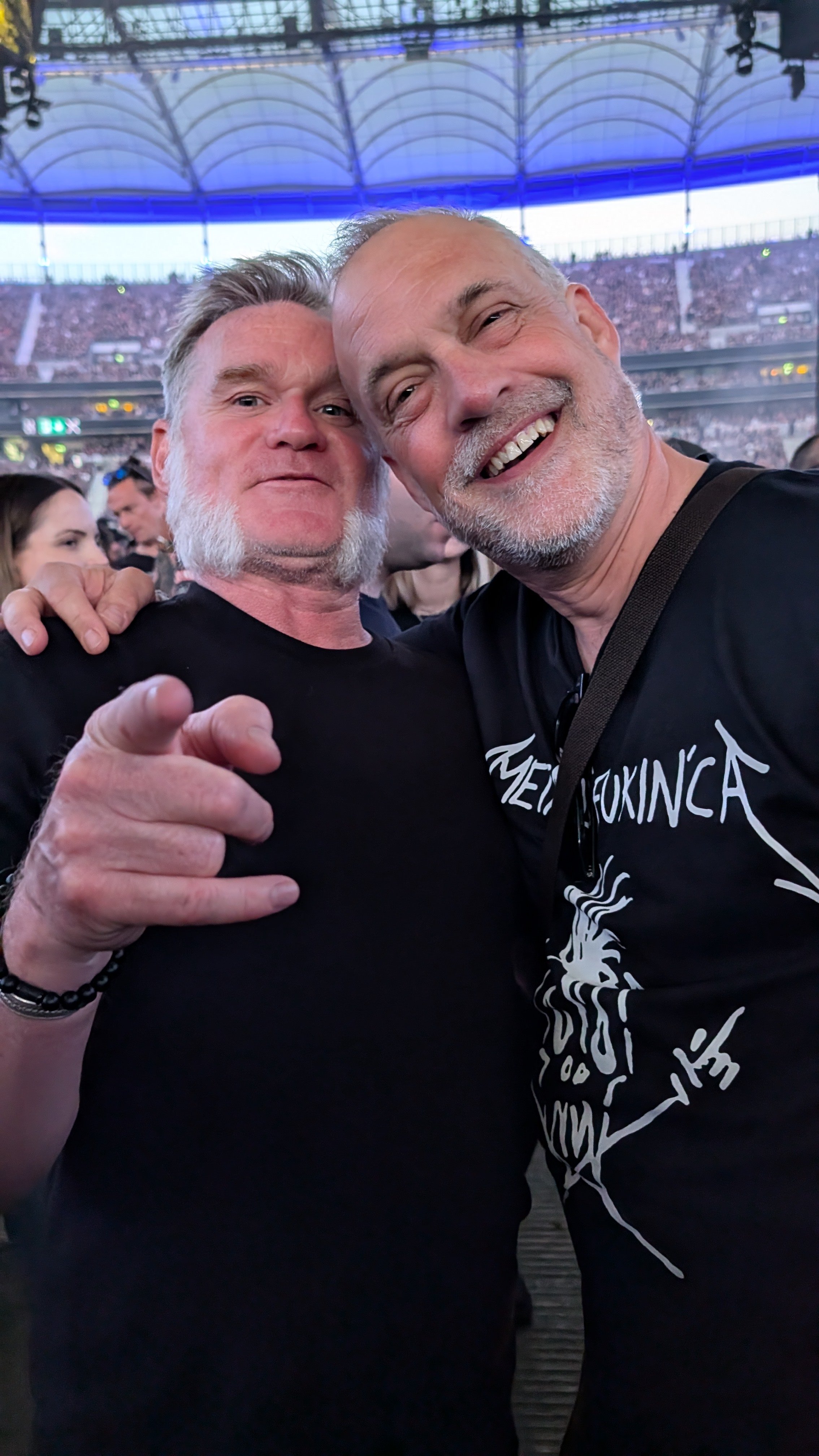 Two men taking a selfie at a concert or stadium, smiling and pointing at the camera, with a crowd and a large roof structure in the background.
