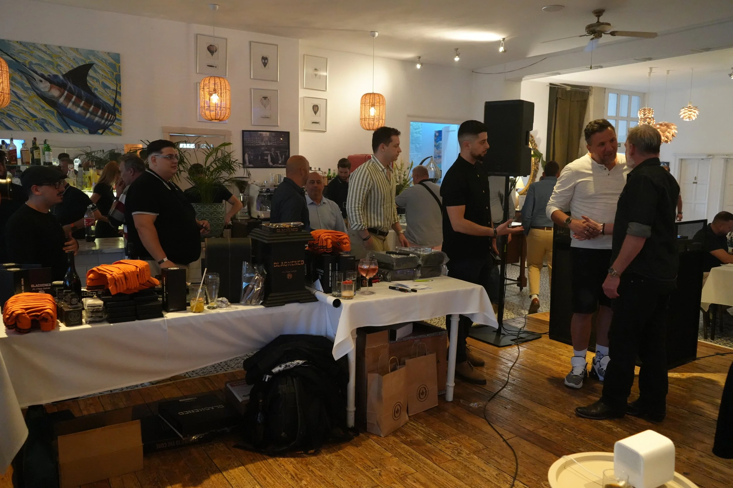 A busy indoor event with several people engaged in conversations. There is a table with orange and black merchandise, possibly for sale, and a bar area with bottles in the background. The room is decorated with artwork and hanging pendant lights, and