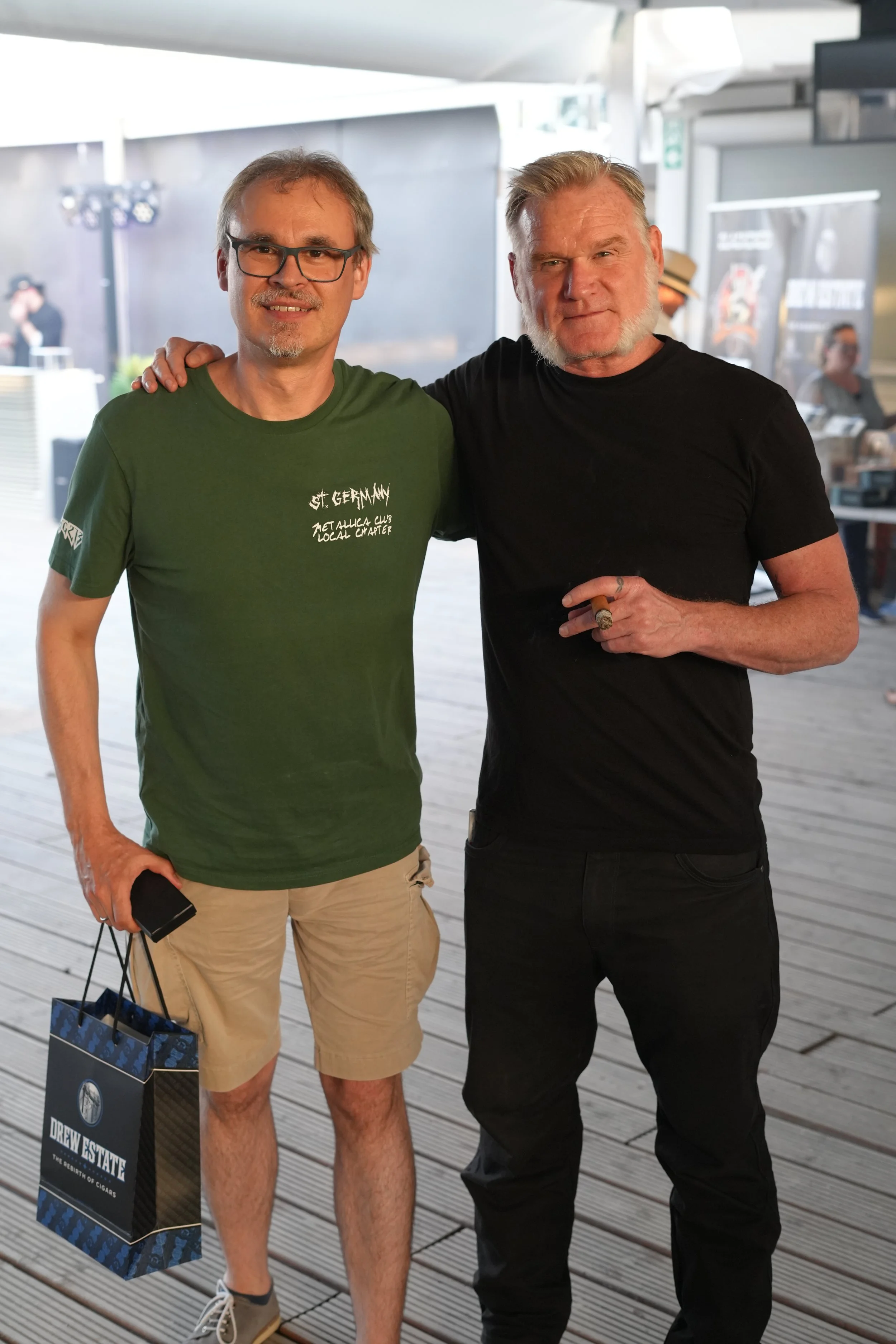 Two men standing outdoors, one wearing glasses, a green t-shirt, and tan shorts holding shopping bags, and the other dressed in black, with a cigar in his hand, both smiling at the camera.