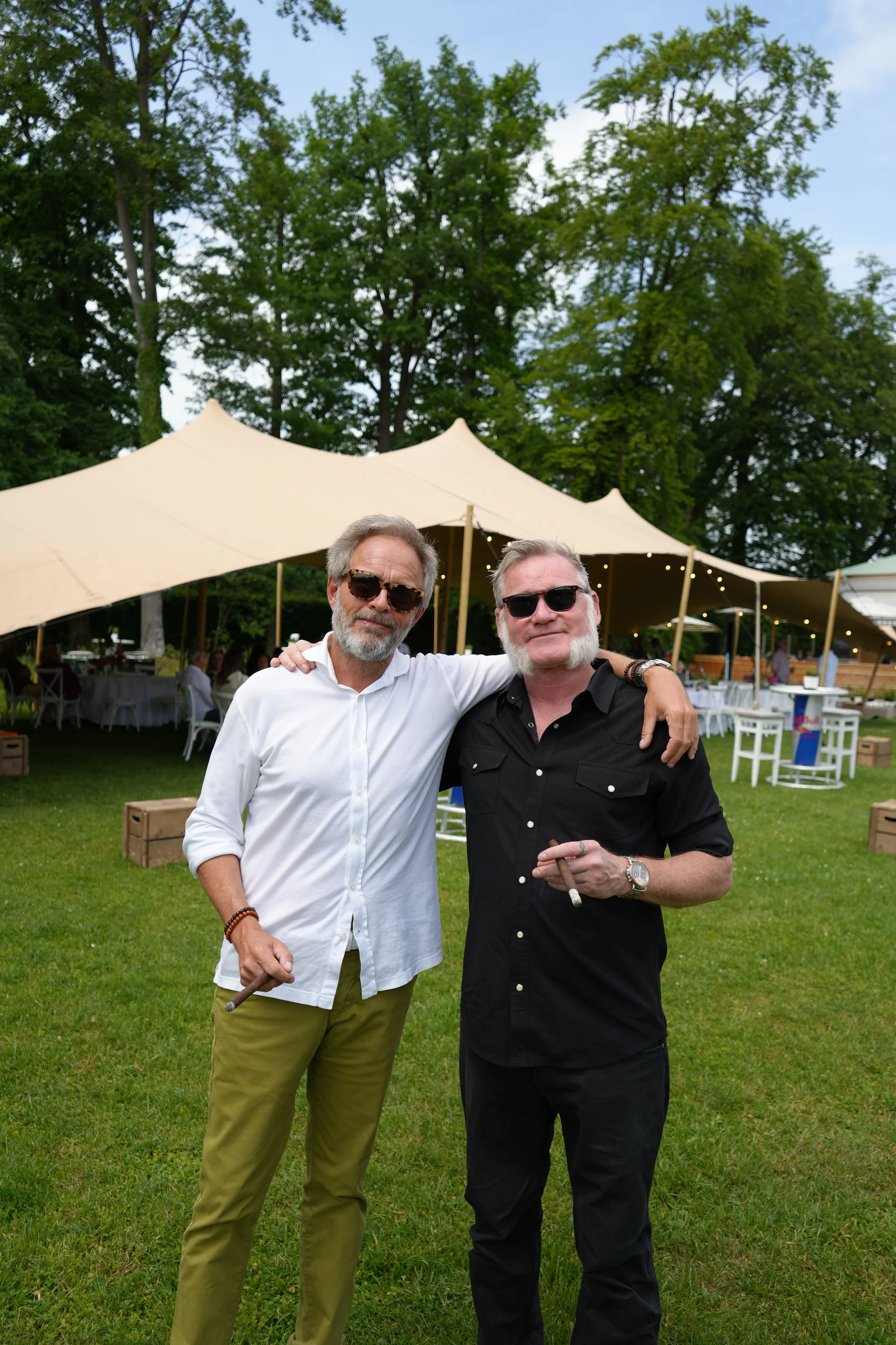 Two middle-aged men with white hair and beards posing outdoors under a large tent at a garden party or outdoor event. One man is wearing a white shirt and the other a black shirt, both with sunglasses, smiling, with trees and tables with white tablec