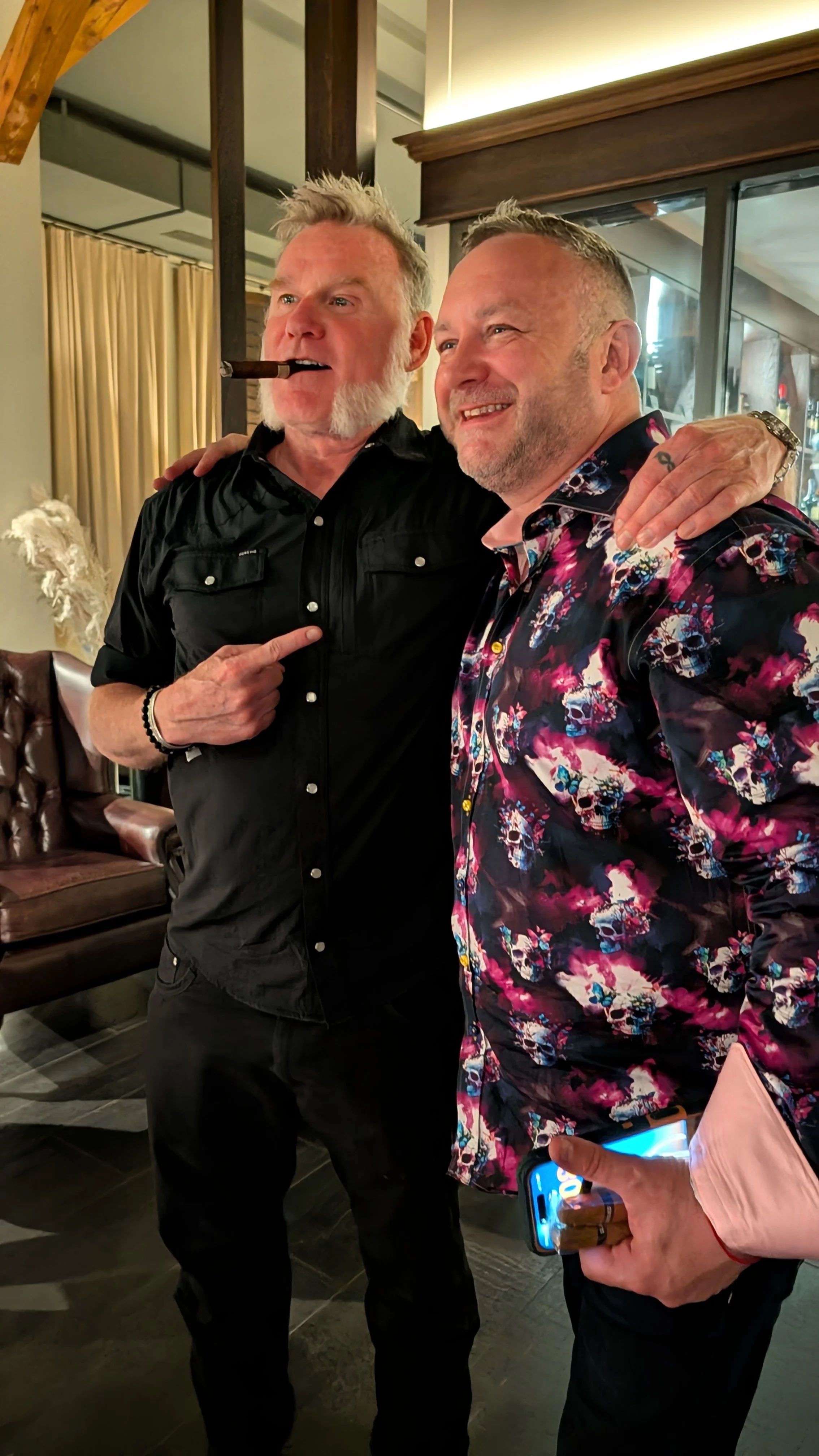 Two men stand close together inside a room with wood accents, one with a cigar in his mouth and the other smiling, both casually dressed, one in a black shirt and the other in a colorful skull-patterned shirt.