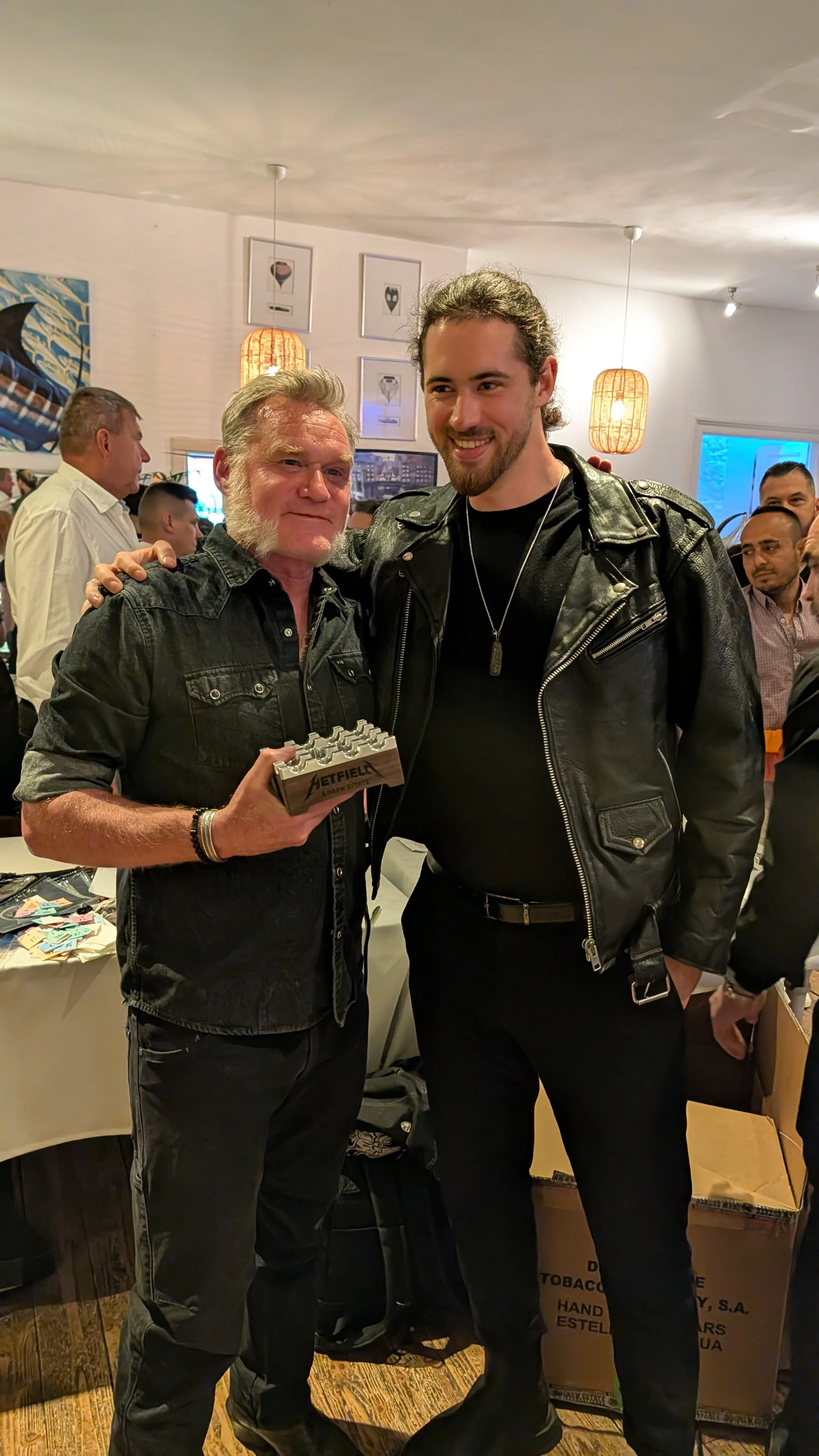 Two men wearing black denim and leather jackets, smiling, standing in a room full of people at an event. One man holds a plaque. Background includes hanging lights, framed art, and a table with items.