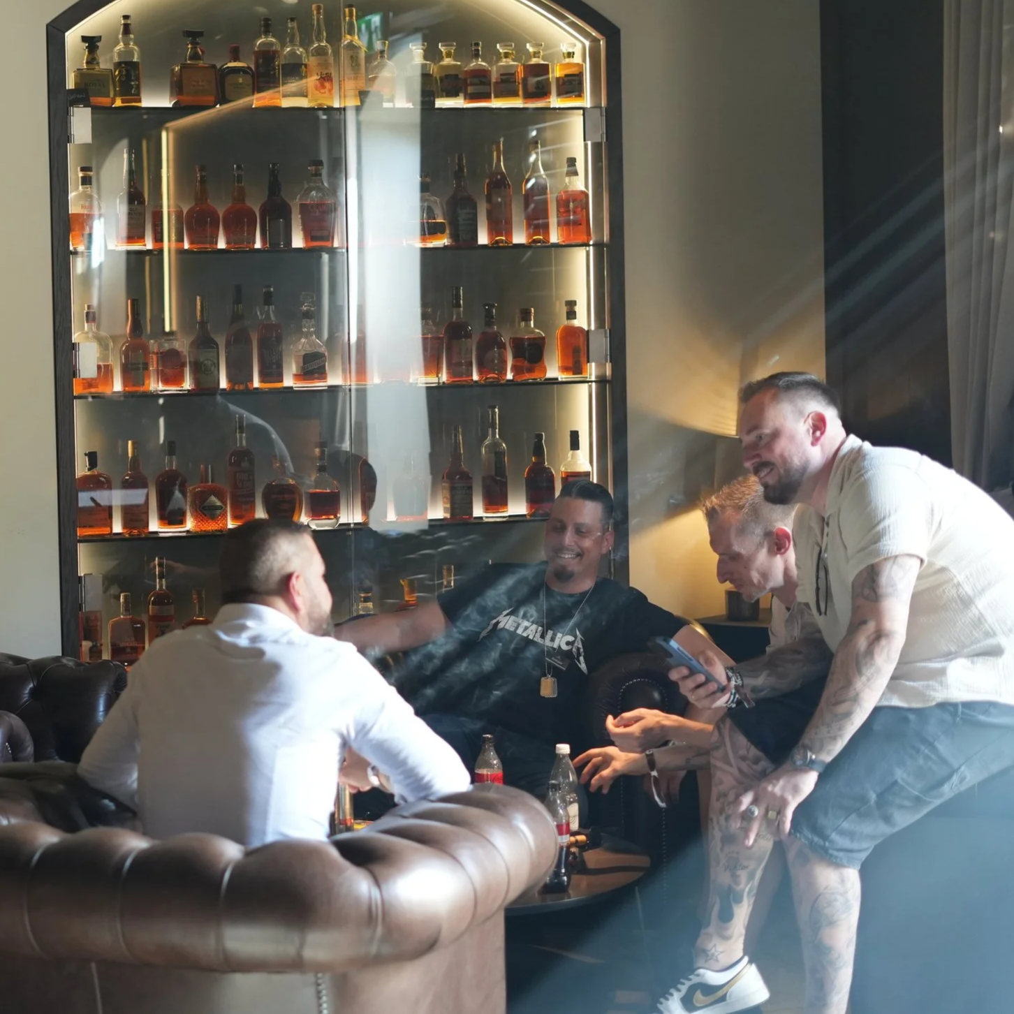Four men are hanging out and having fun in a lounge or bar with a large display of liquor bottles behind them. They are smiling and talking, and some are holding drinks.