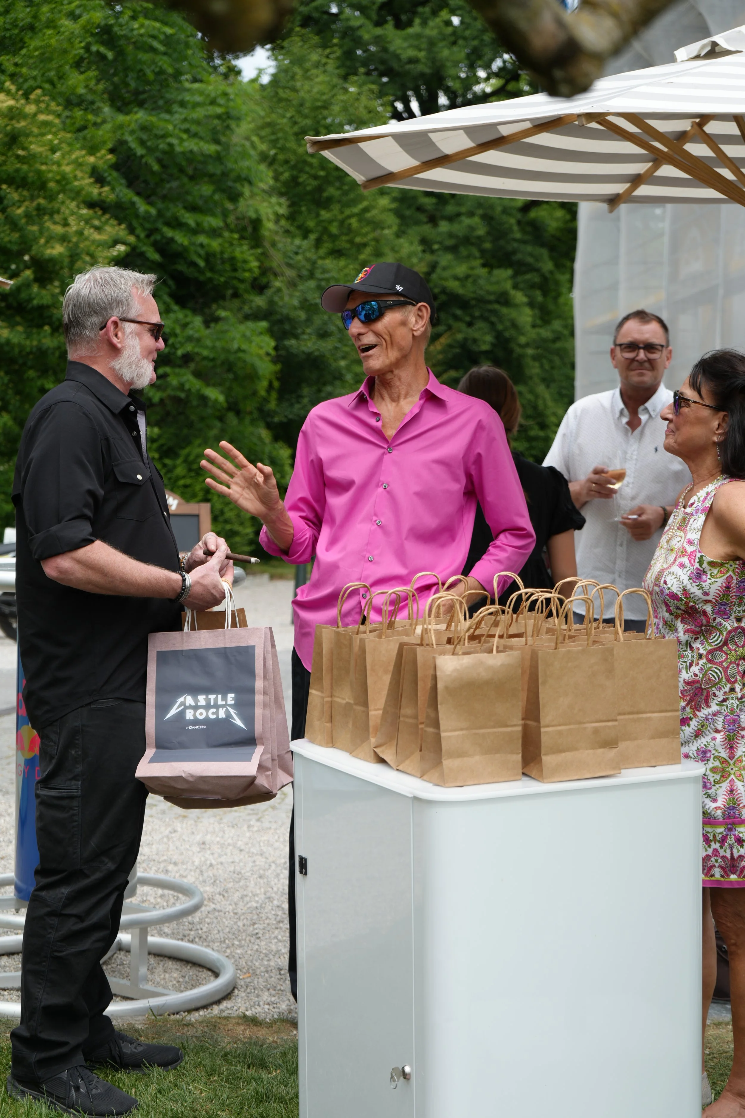 People gathered outdoors around a table with brown paper bags, under a large white and gray striped umbrella, with trees and a building in the background.