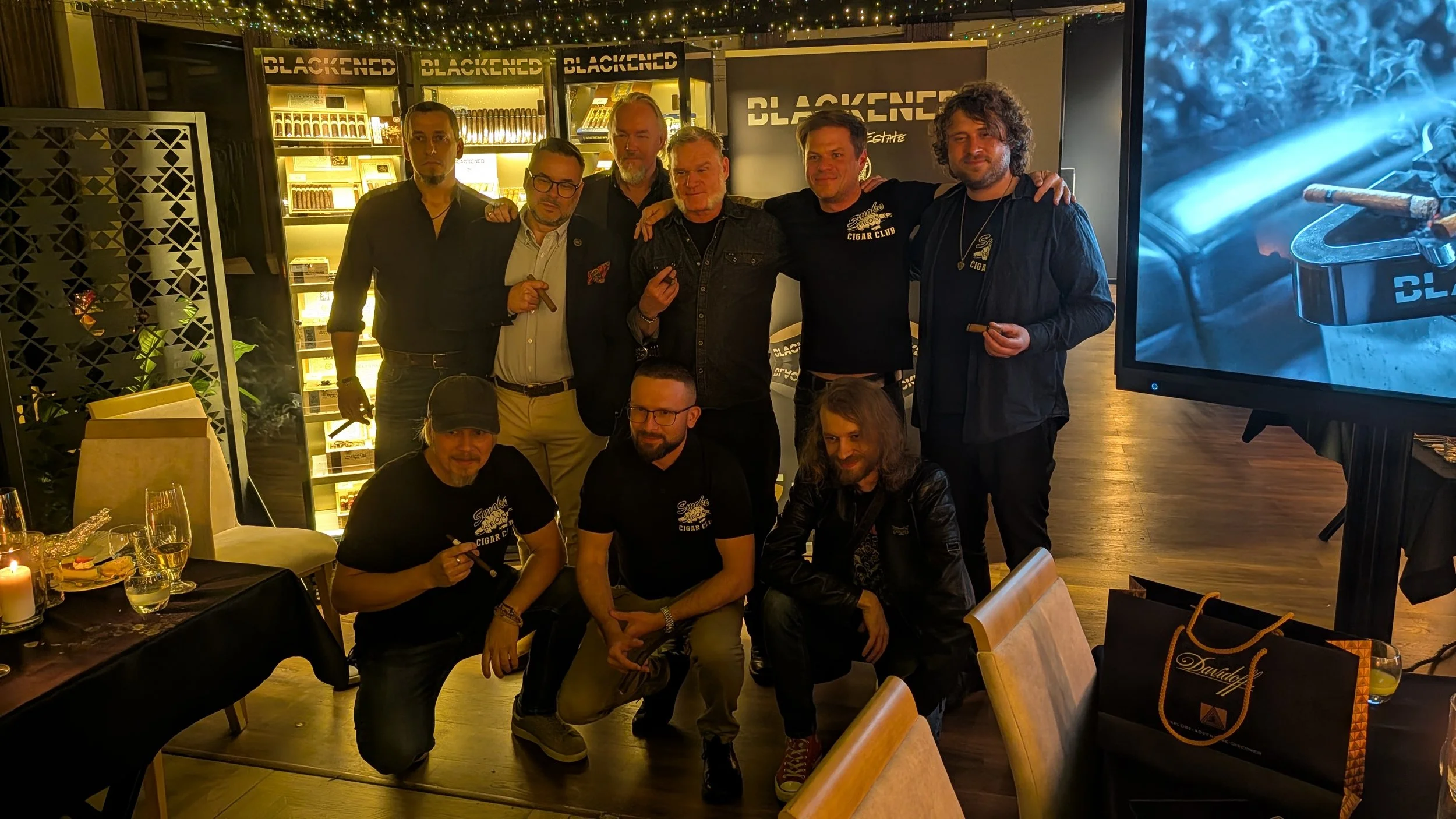 Group of ten men posing together at a cigar event, with a display of cigars behind them and a lit cigar with blue smoke in the foreground.