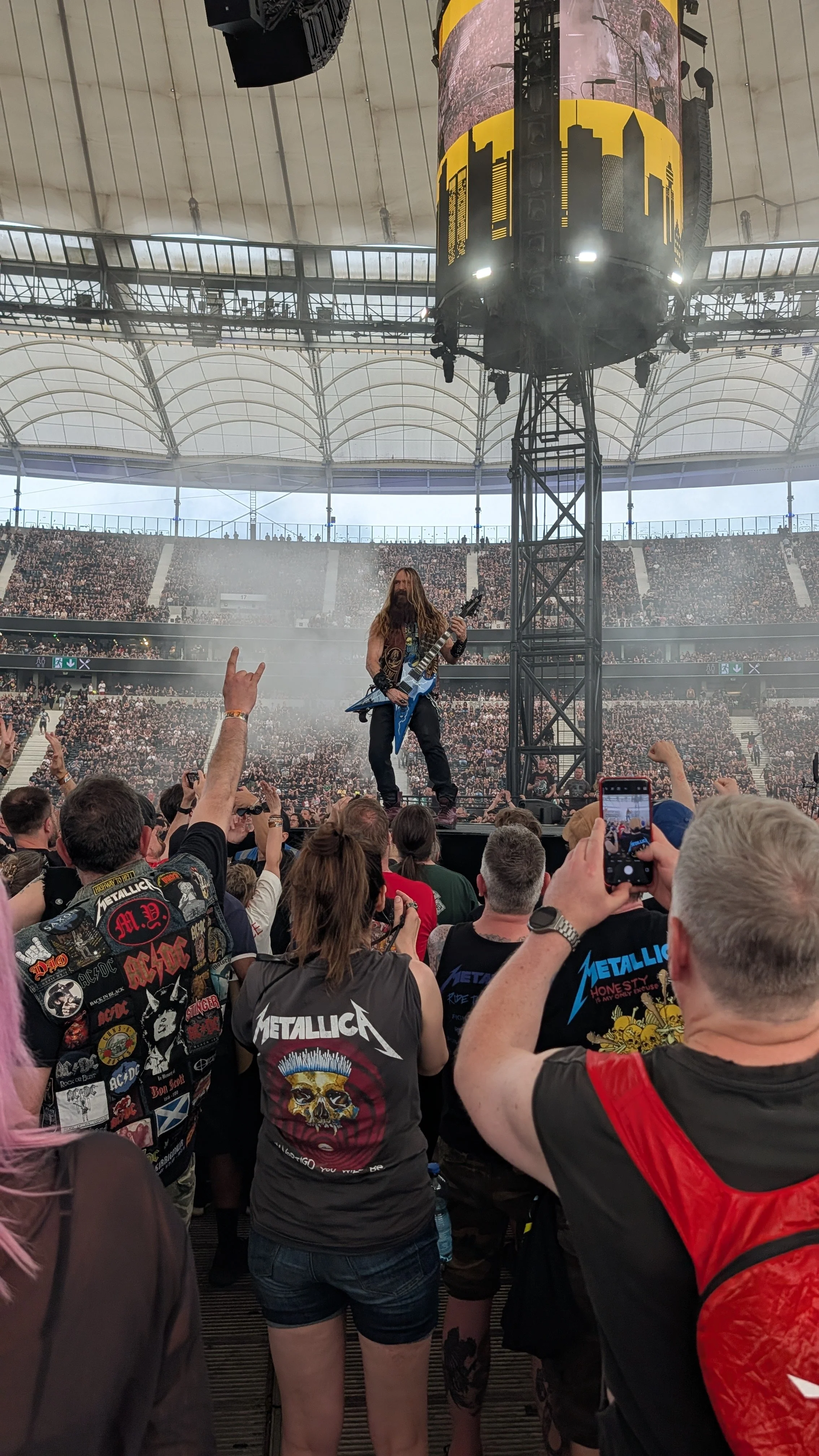 A musician with long hair and a black sleeveless shirt is playing an electric guitar on stage at a concert, with a large crowd in the background. The stage features a tall scaffolding structure and a large screen displaying a city skyline silhouette 