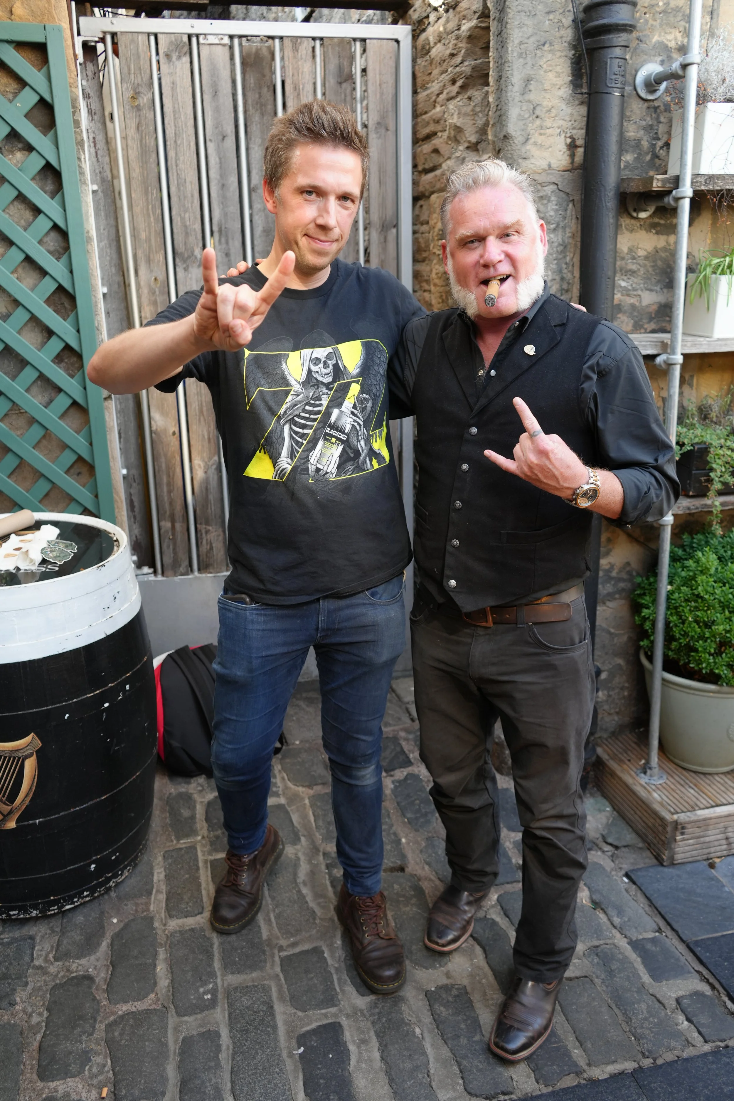 Two men standing outdoors on a cobblestone patio, smiling and making hand gestures. One man has brown hair, wearing a black T-shirt with a skeleton graphic and is making the horns gesture. The other man has gray hair, a beard, a cigar in his mouth, a
