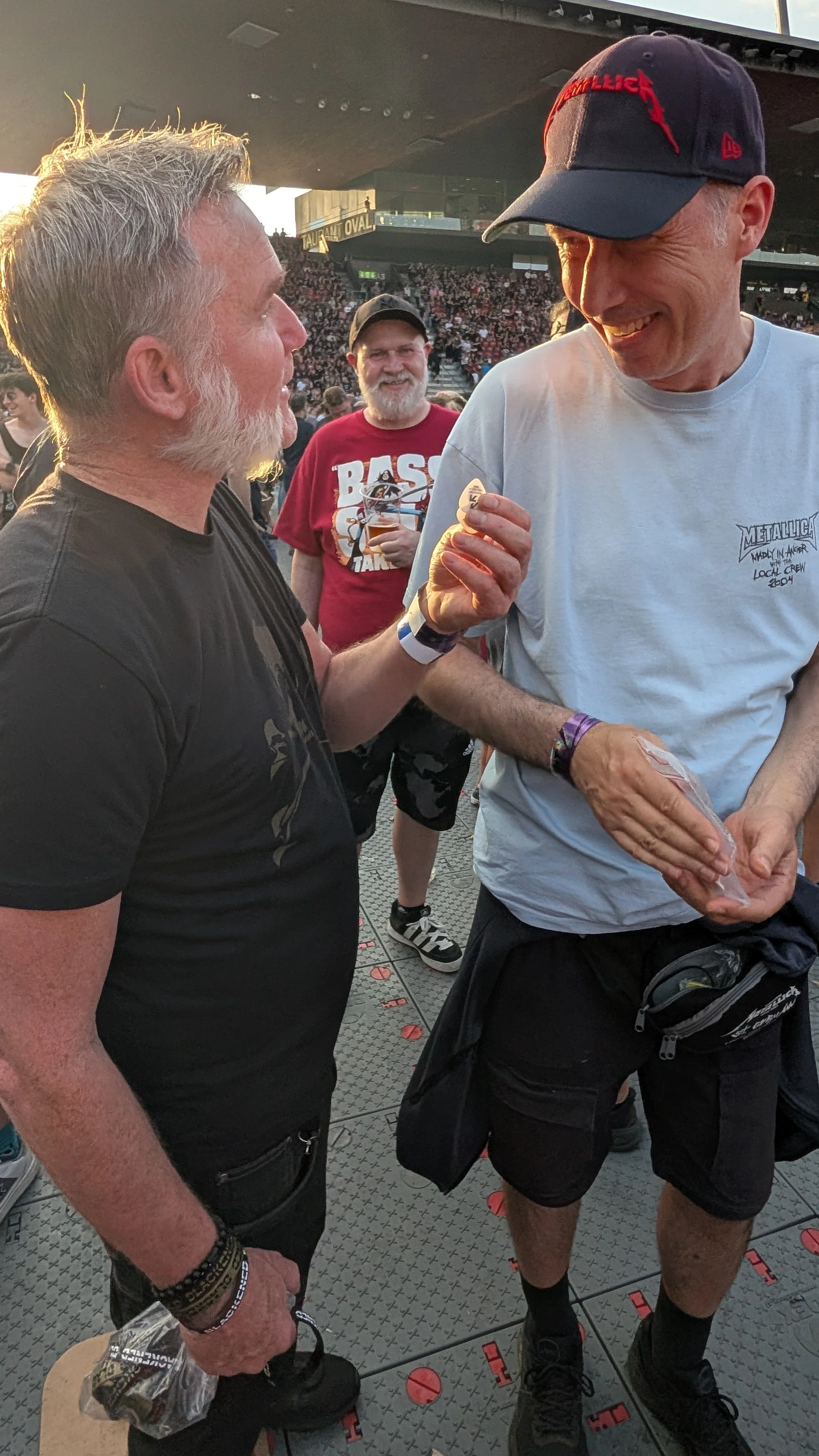 Two men are engaged in a conversation at a Metallica concert, with a third person observing in the background. They are both smiling, and the man on the right is holding a plastic bag and a guitar picks.