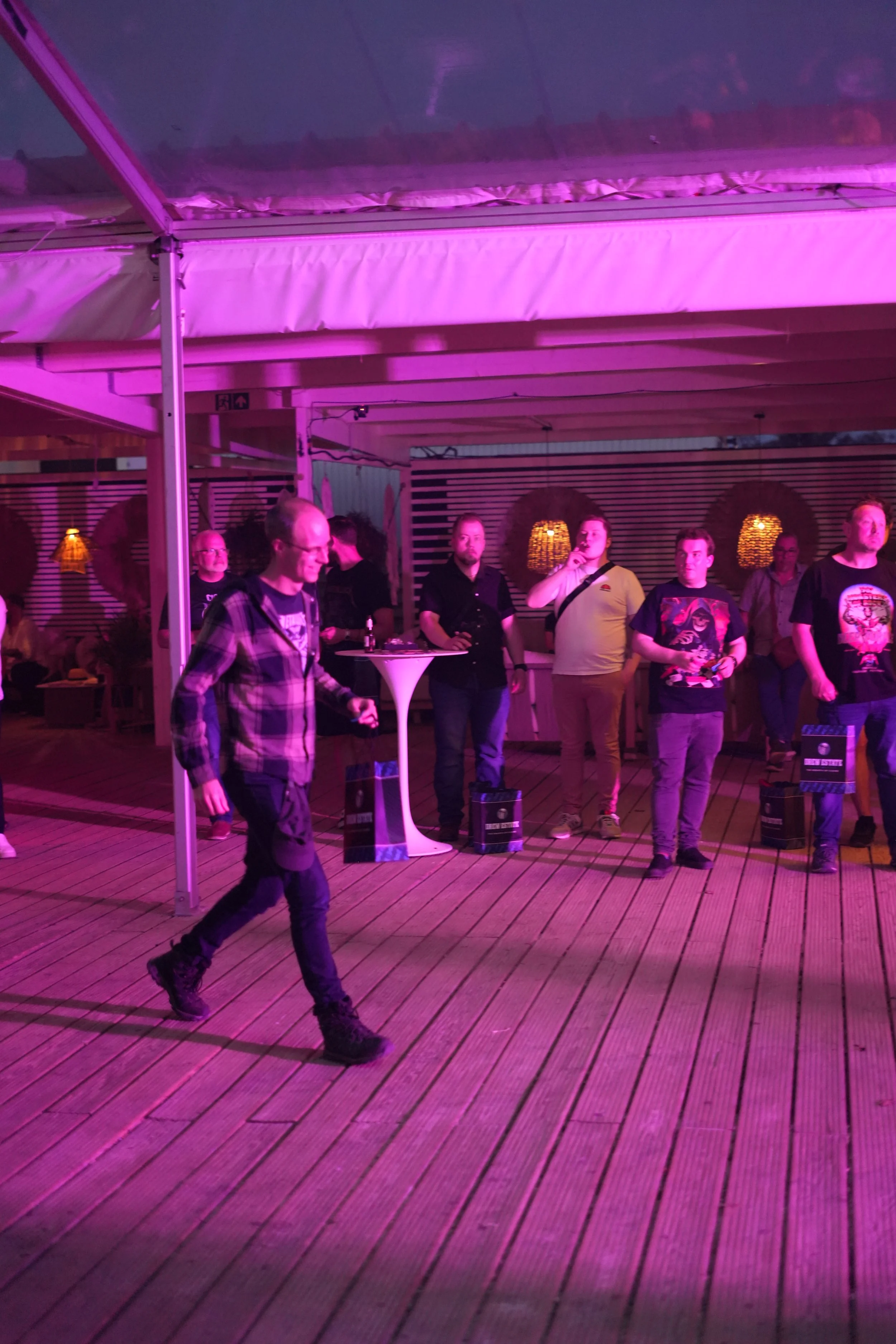 People attending a social event under purple lighting inside a wooden-floored venue, some standing and one person walking in the foreground.