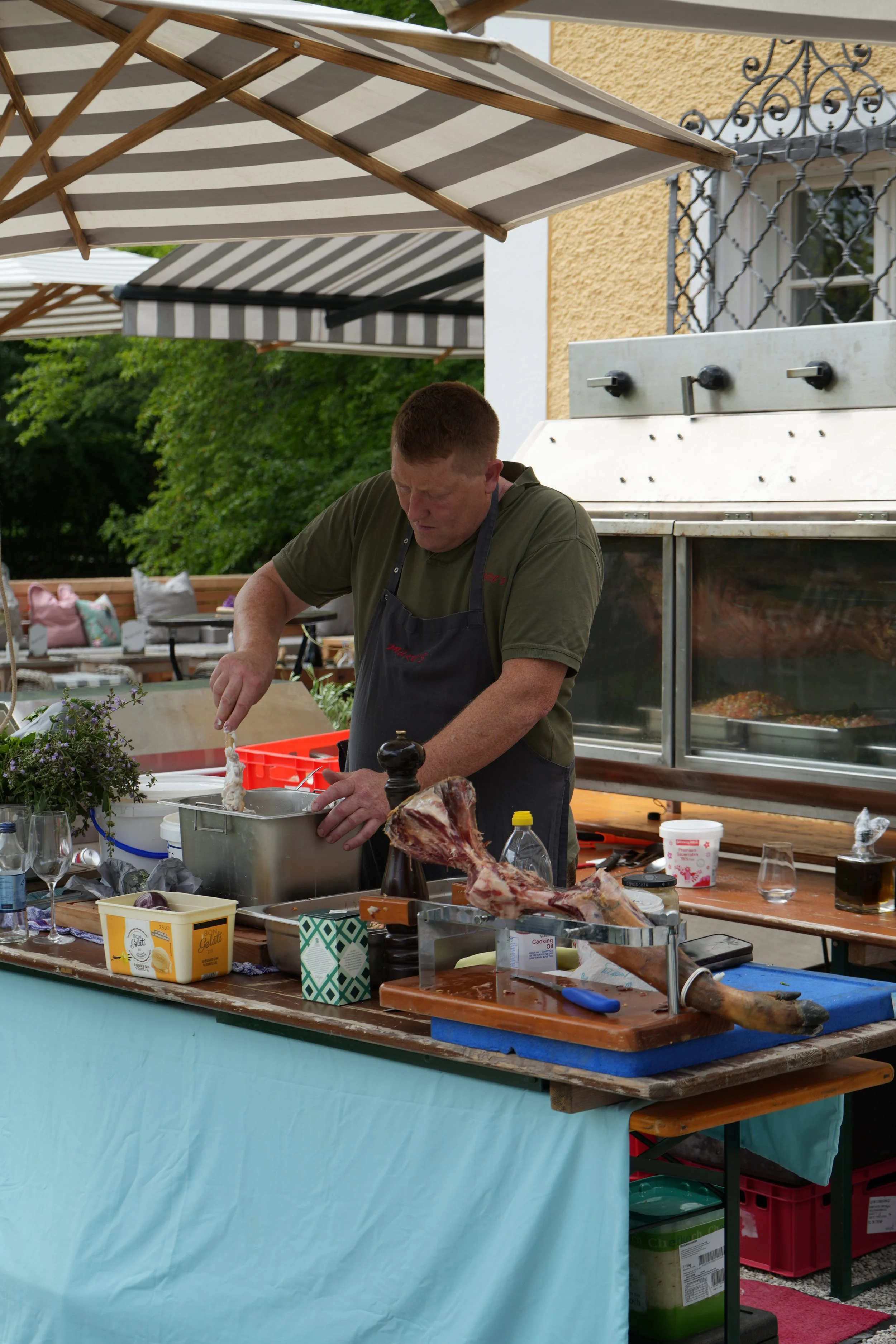 A man preparing food at an outdoor barbecue station, with a large piece of raw meat on a cutting board, various cooking supplies, and a grill in the background under striped umbrellas.