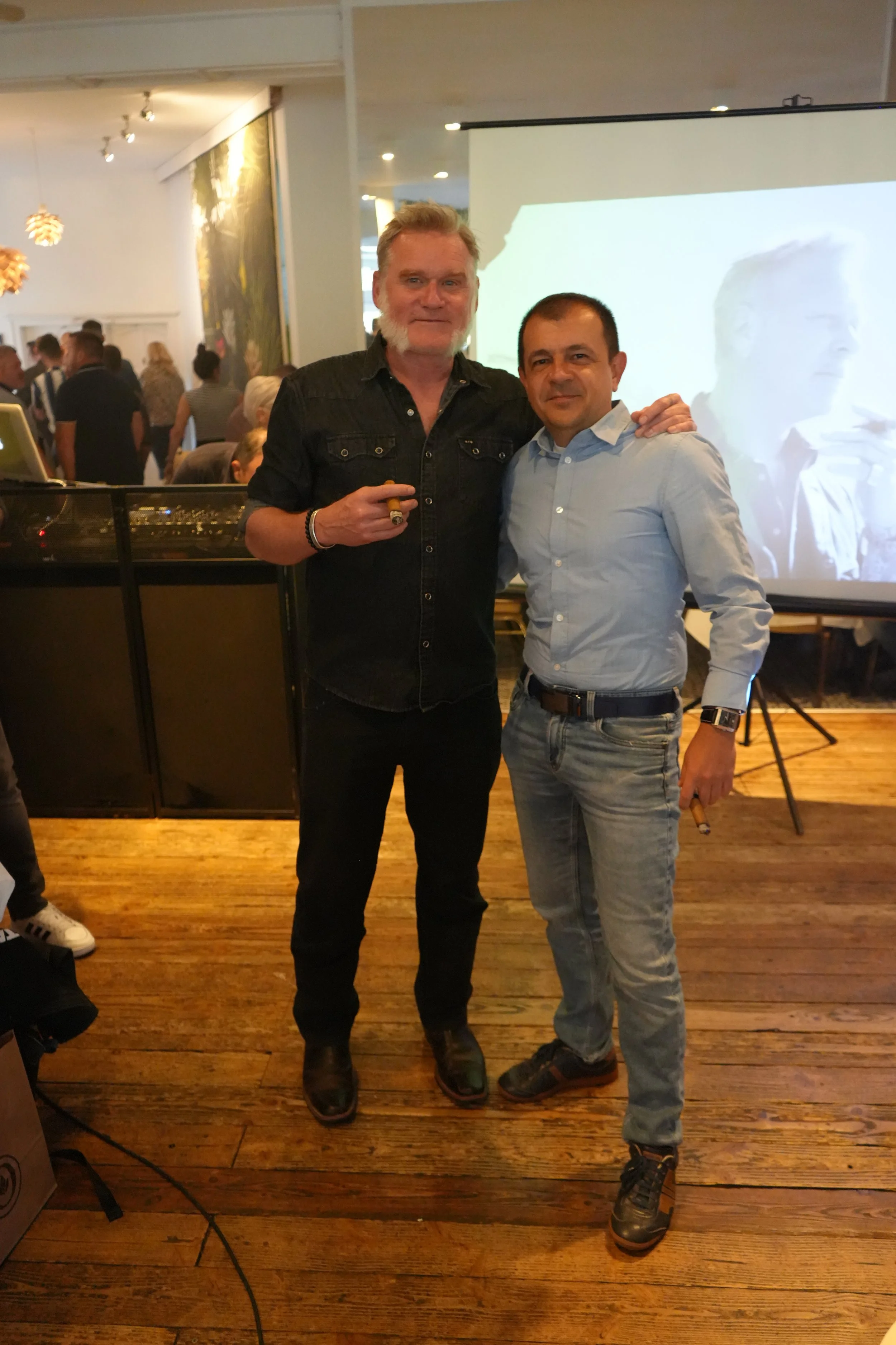 Two men standing together at an indoor event, with a large screen behind them and people in the background. They are smiling, one has gray hair and the other has dark hair, both dressed casually.