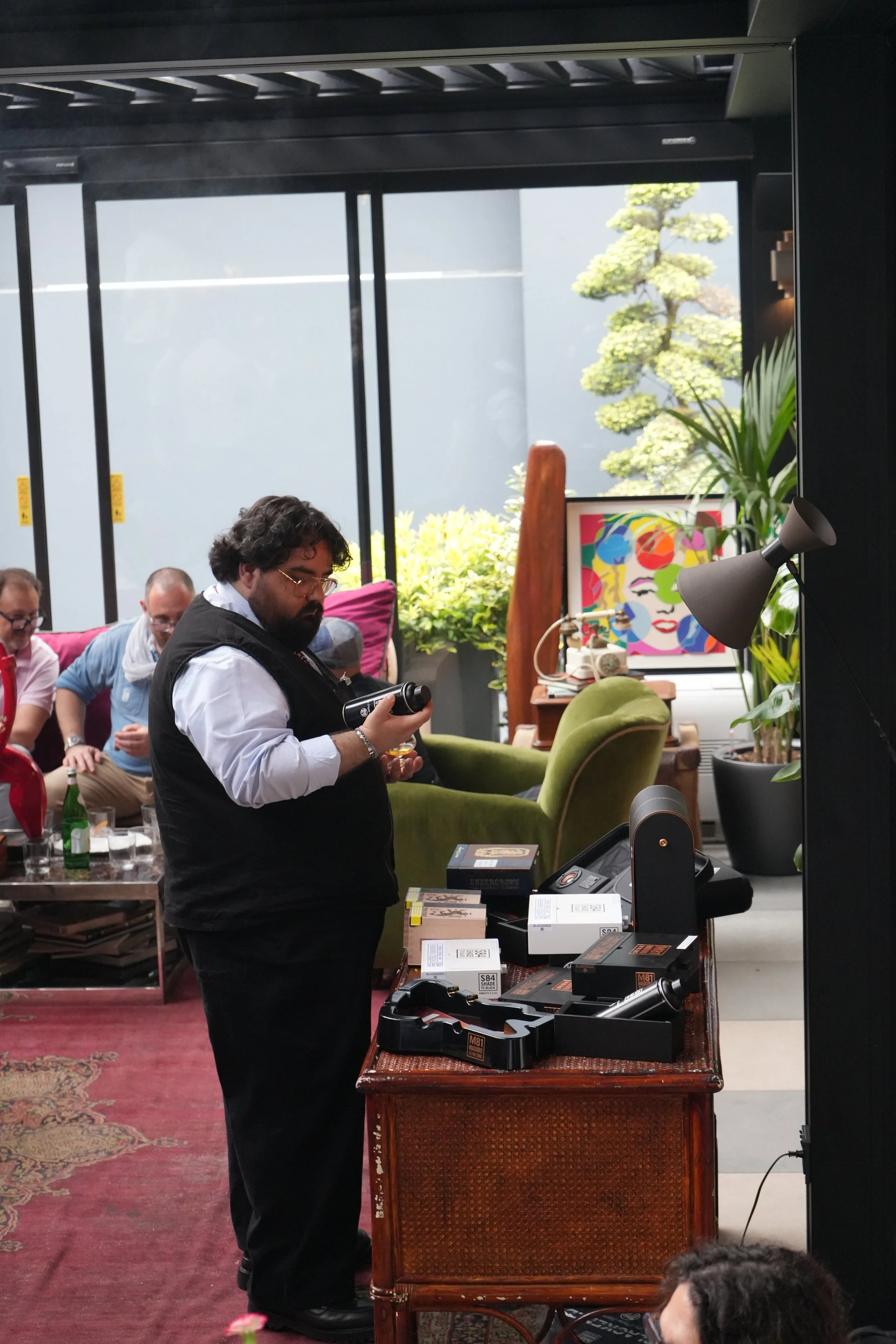 A man with dark curly hair and glasses, dressed in a white shirt and black vest, is standing at a table displaying various boxed camera equipment. Behind him is a colorful portrait art piece of a woman's face, greenery and a tree outside through larg