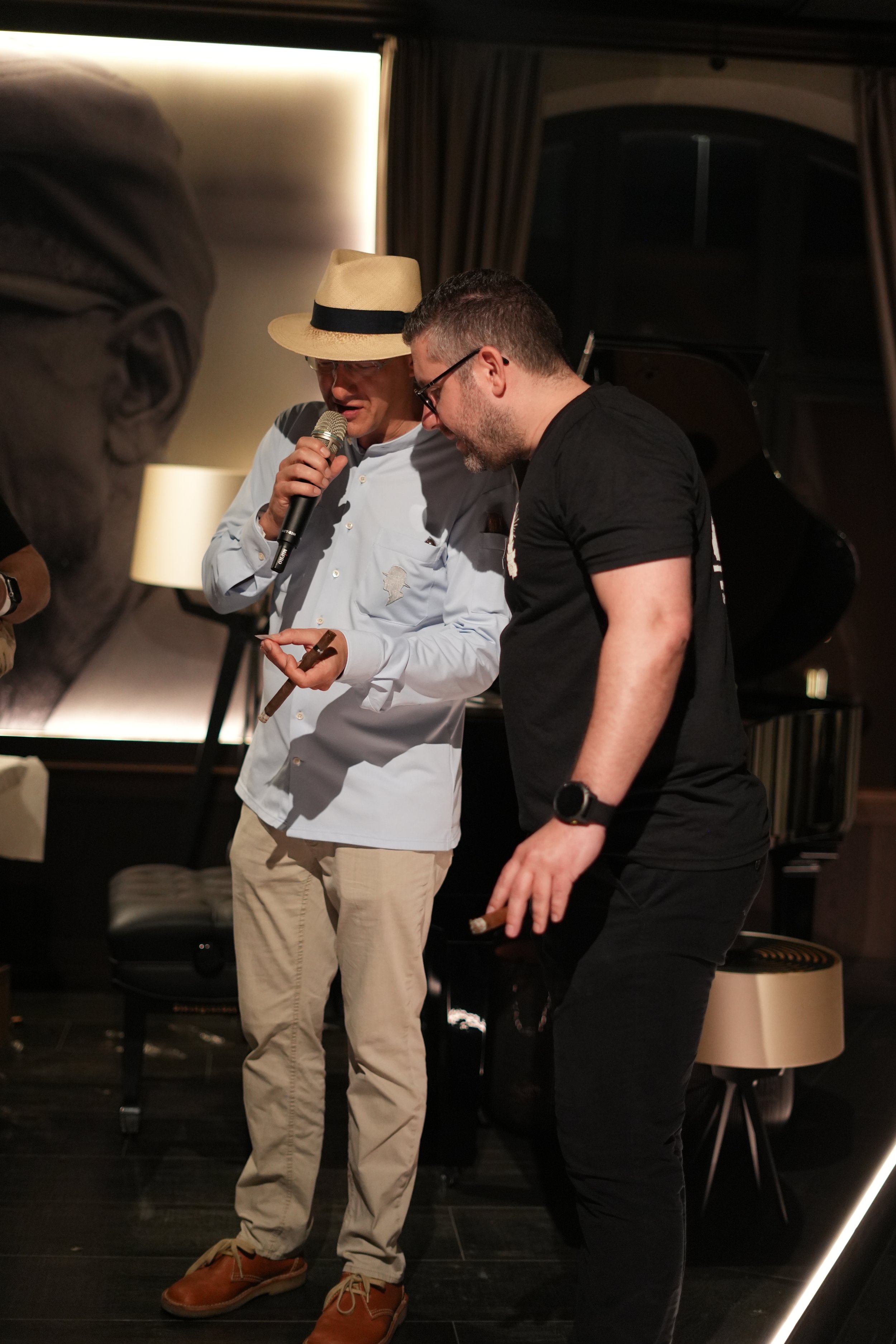 Two men with glasses and a cigar and a microphone and notepad are standing close together inside a room with dark furniture. One man is wearing a straw hat and white shirt, the other is wearing a black T-shirt. They are looking at each other and seem