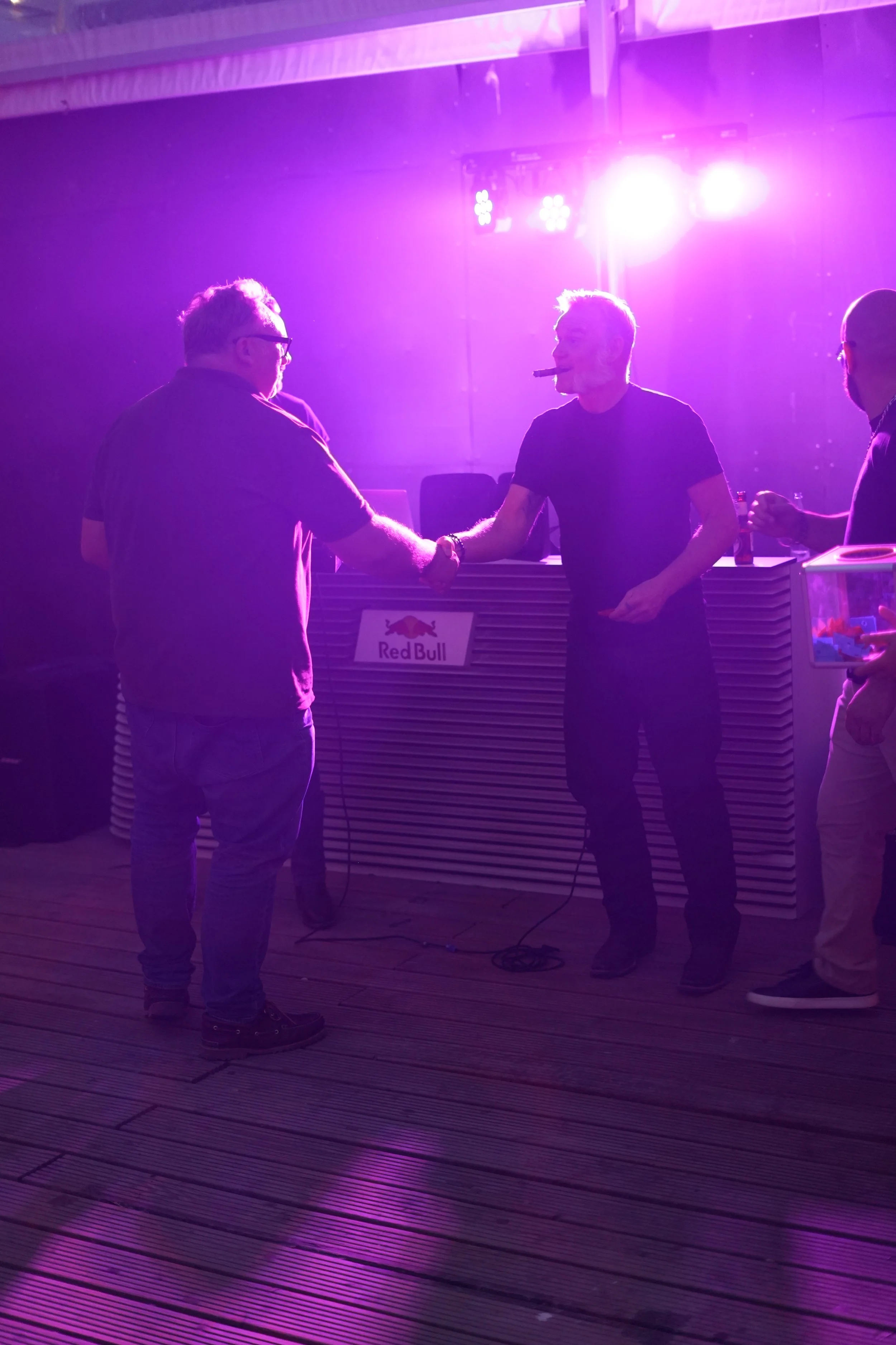 People at a dimly lit party or event with purple lighting, one person on the right smoking a cigar, and a bar with the Red Bull logo in the background.