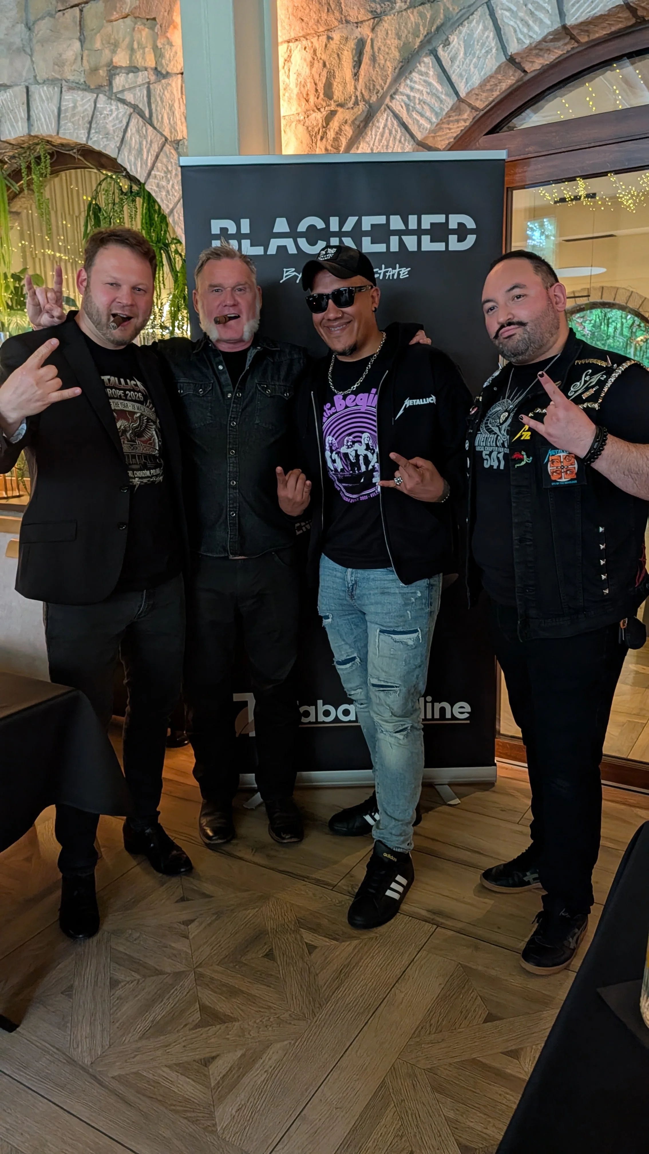 Four men posing in front of a sign that reads 'BLACKEND' at an indoor event, with all four making rock and roll hand gestures and smiling.