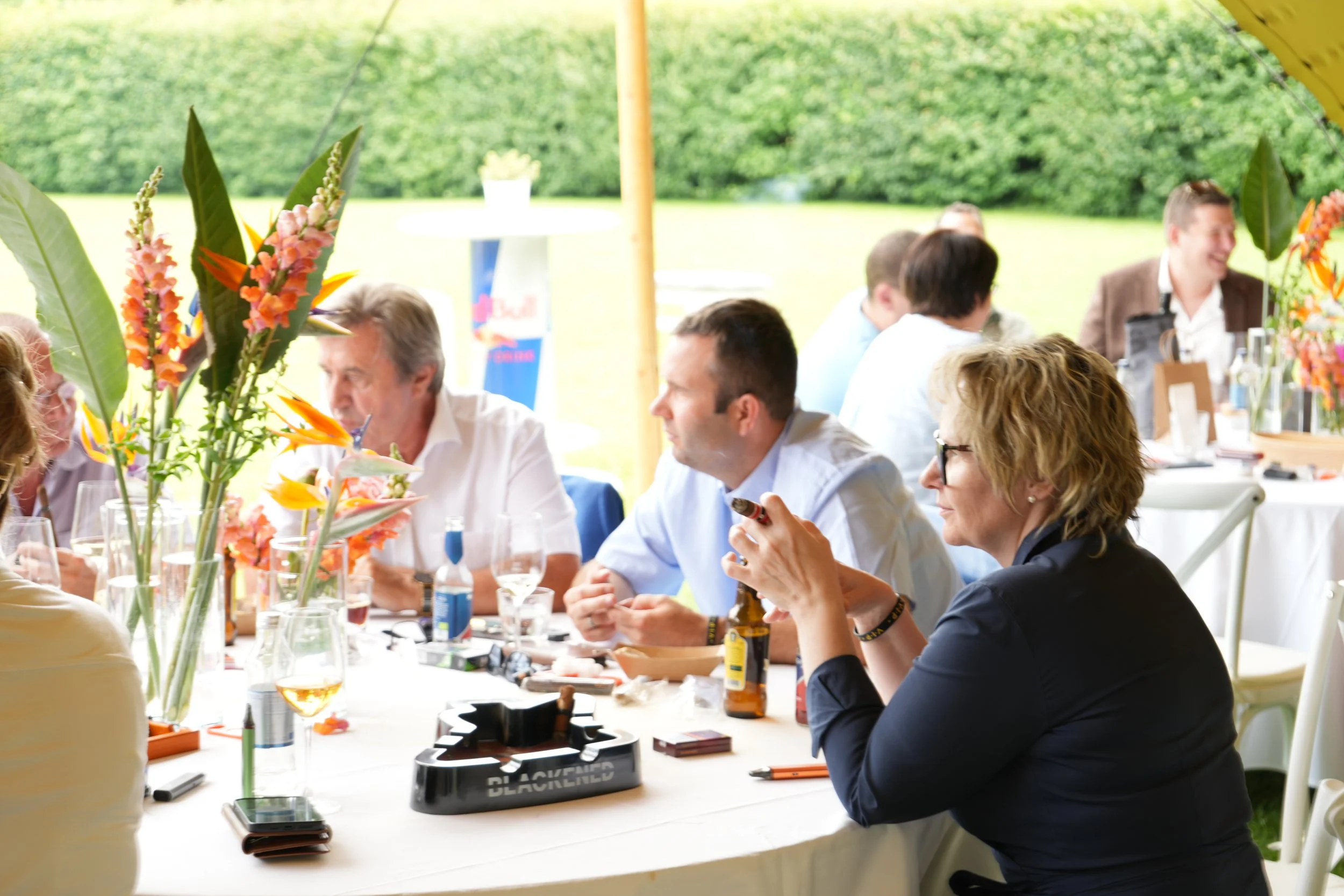People sitting at a long table under a tent outdoors, engaging in conversation, with floral centerpieces and drinks on the table.