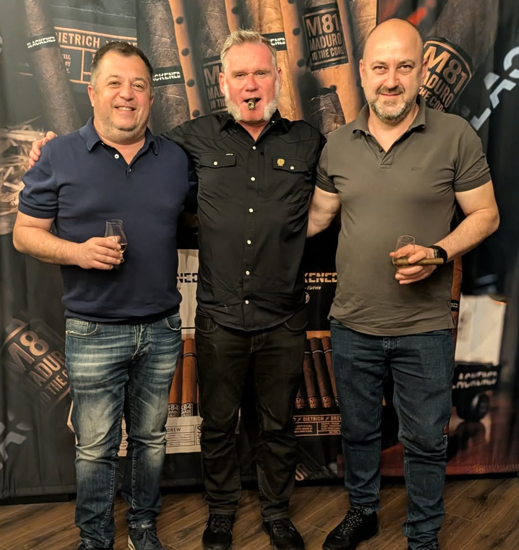 Three men standing together, smiling, in front of a backdrop with cigar images and radio station logos. The man on the left is holding a glass with a drink, wearing a navy blue polo and jeans. The man in the middle is smoking a cigar, wearing a black