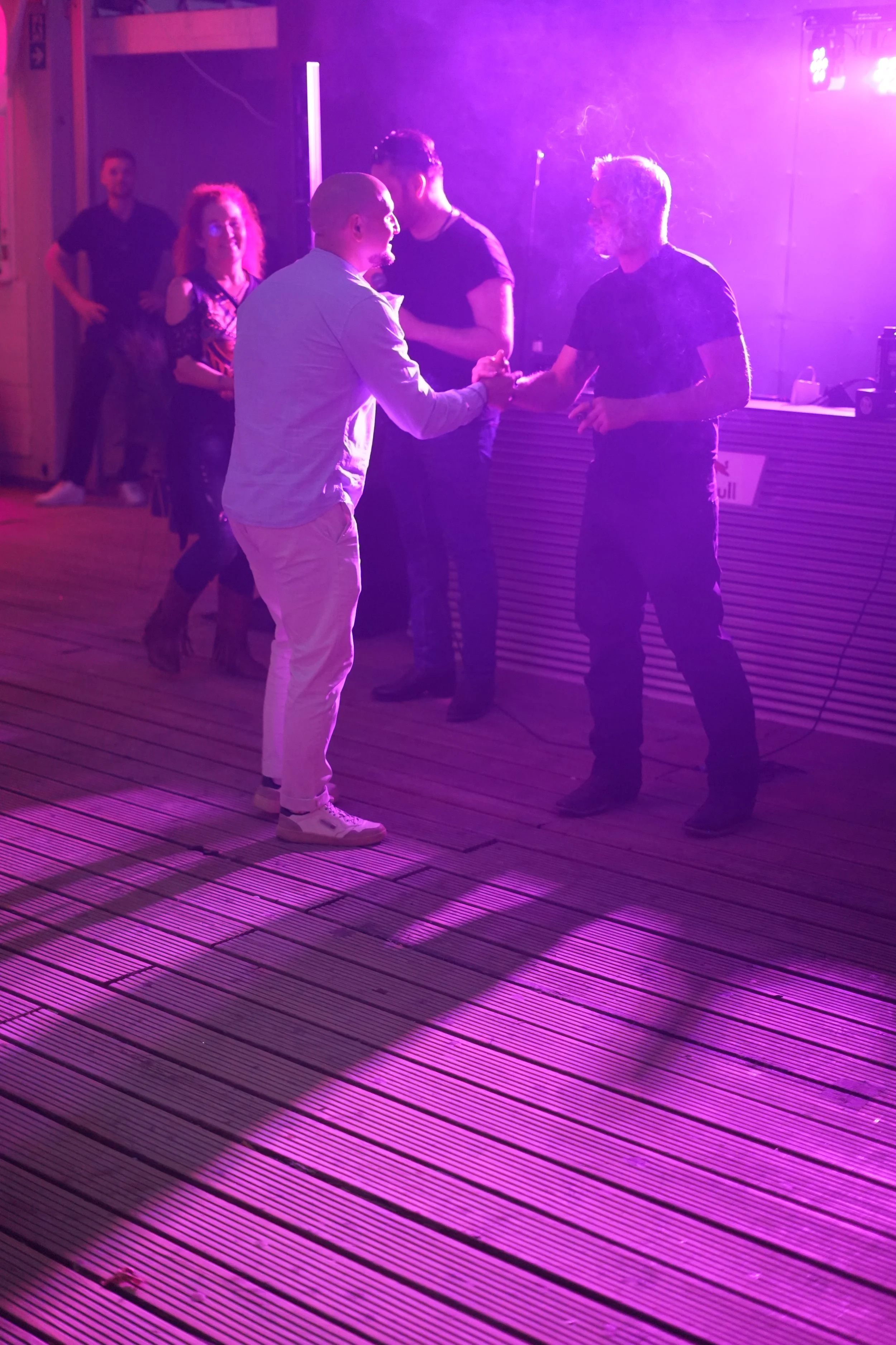 Two men shake hands in a dimly lit nightclub or bar with colorful purple and pink lighting, while three other people watch in the background.