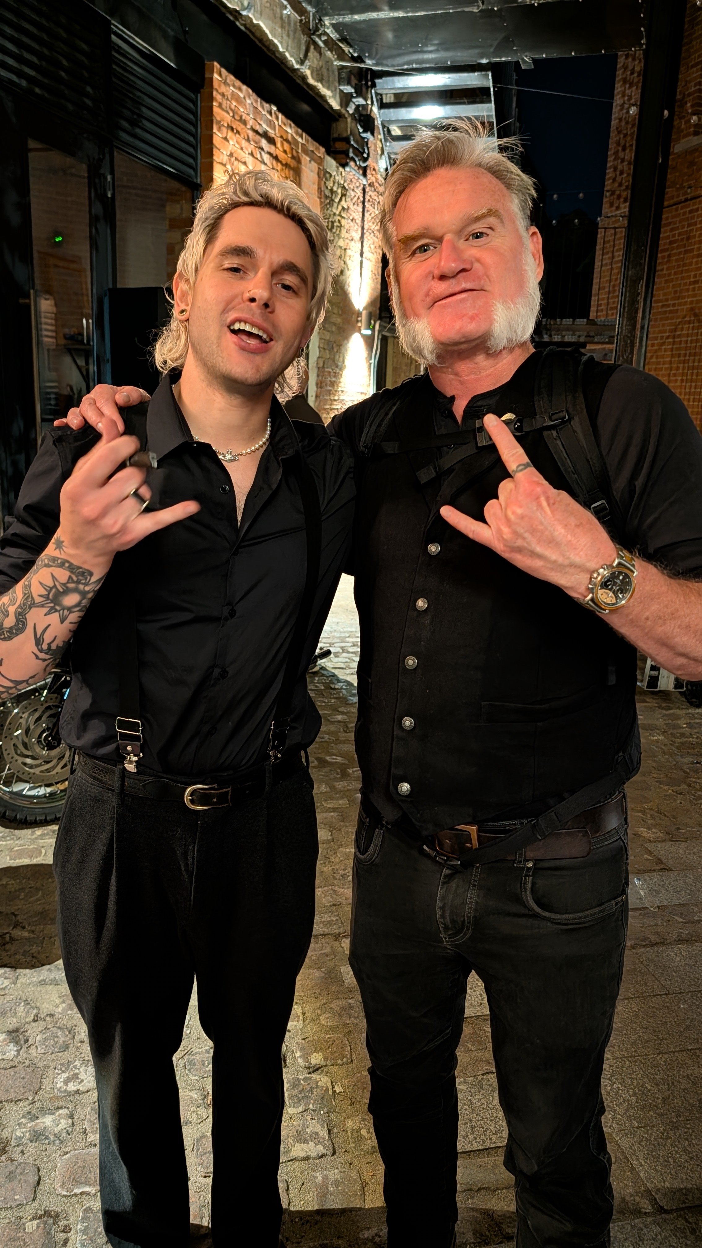 Two men standing arm in arm at night outside, both wearing black shirts and making rock hand gestures. They have light-colored hair, one with a beard and mustache, and the other with short hair and tattoos on his arm.