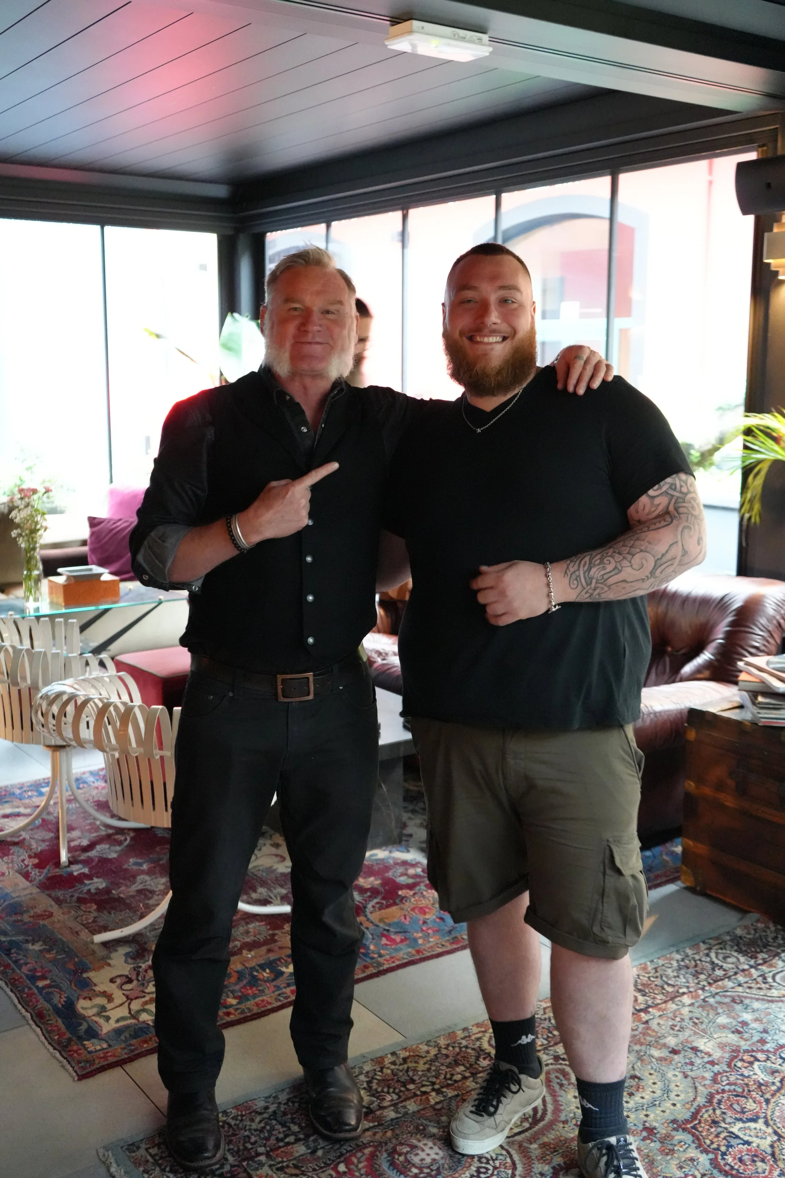 Two men standing inside a room with large windows. One man has long gray hair and a beard, wearing a black shirt and pointing at the other man. The other man has a beard, tattoos on his arm, wearing a black t-shirt, shorts, and sneakers. They are smi