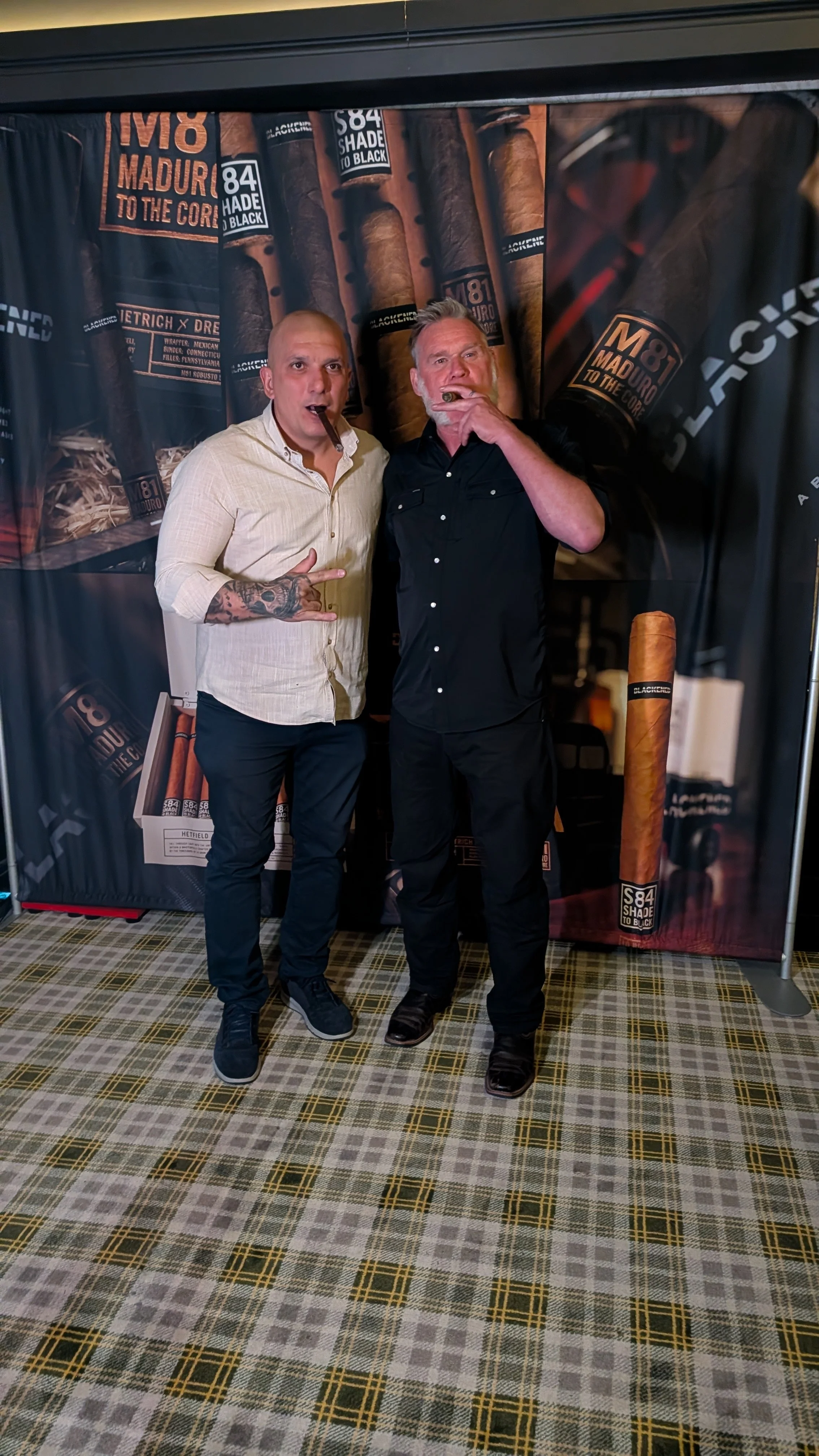 Two men standing in front of a cigar-themed backdrop, smoking cigars and making hand gestures. One man is wearing a light-colored shirt and the other is in black. They are on a checkered carpeted floor.