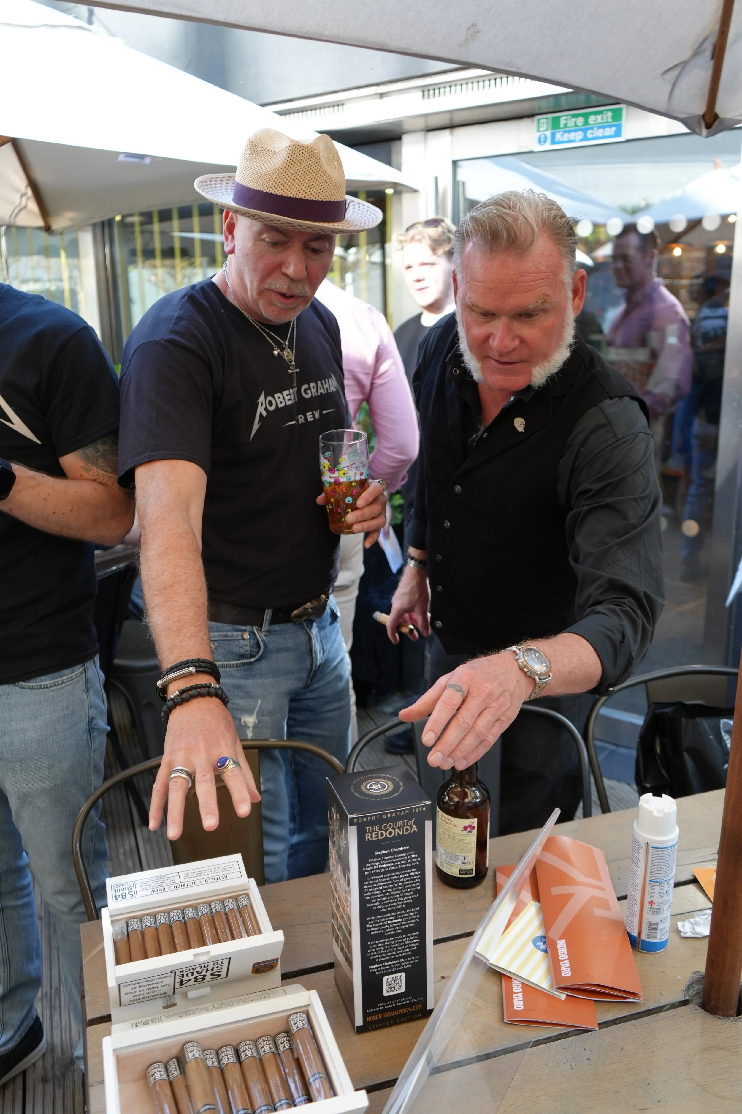 Two men are standing at a table with various cigar boxes and bottles, one wearing a straw hat and the other with white hair and a beard, engaged in conversation or a transaction outdoors.