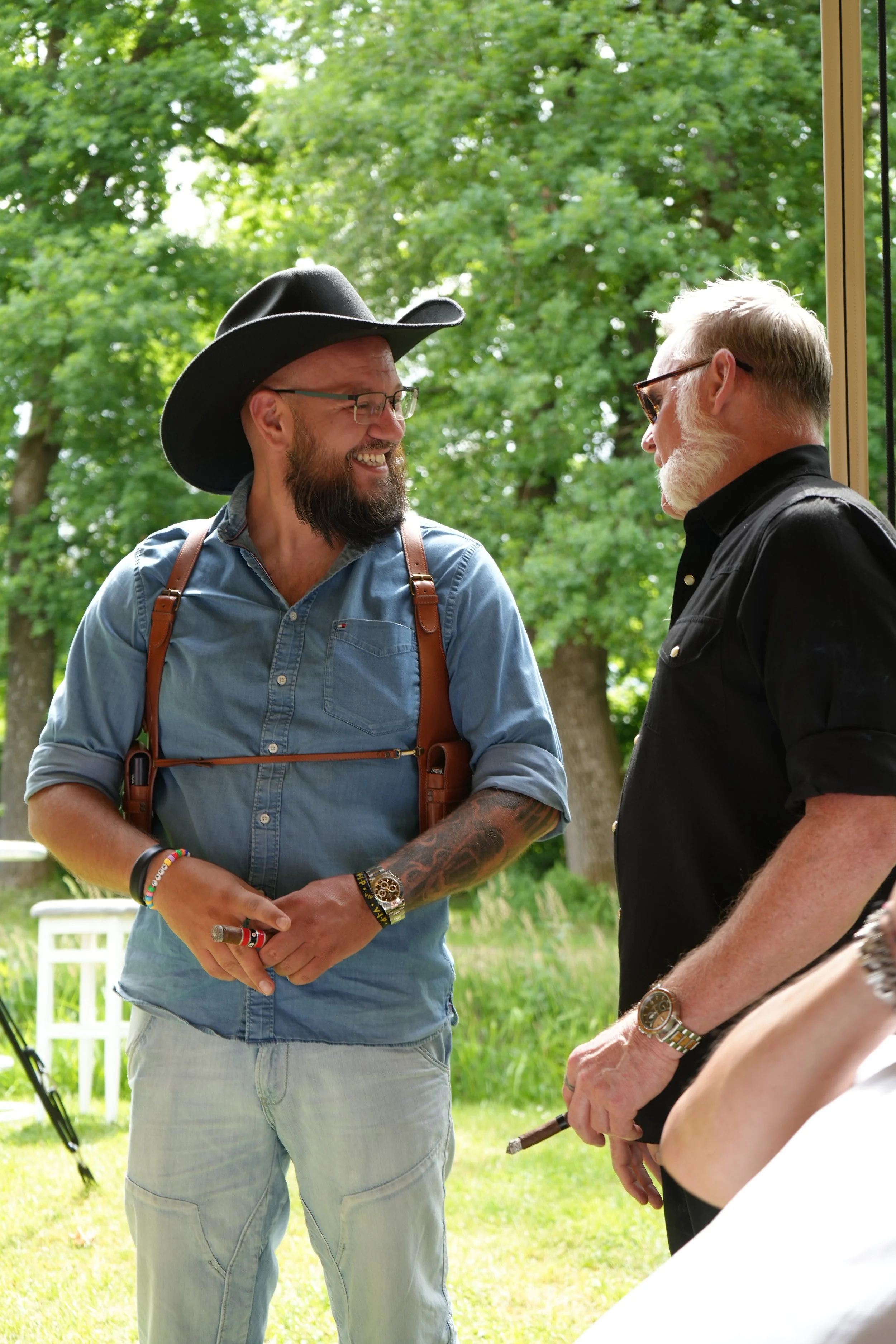 Two men are smiling and engage in conversation outdoors. One man is wearing a black cowboy hat, glasses, a denim shirt, and light-colored jeans, with various bracelets and a tattooed arm. The other man, with gray hair and beard, sunglasses, and a bla