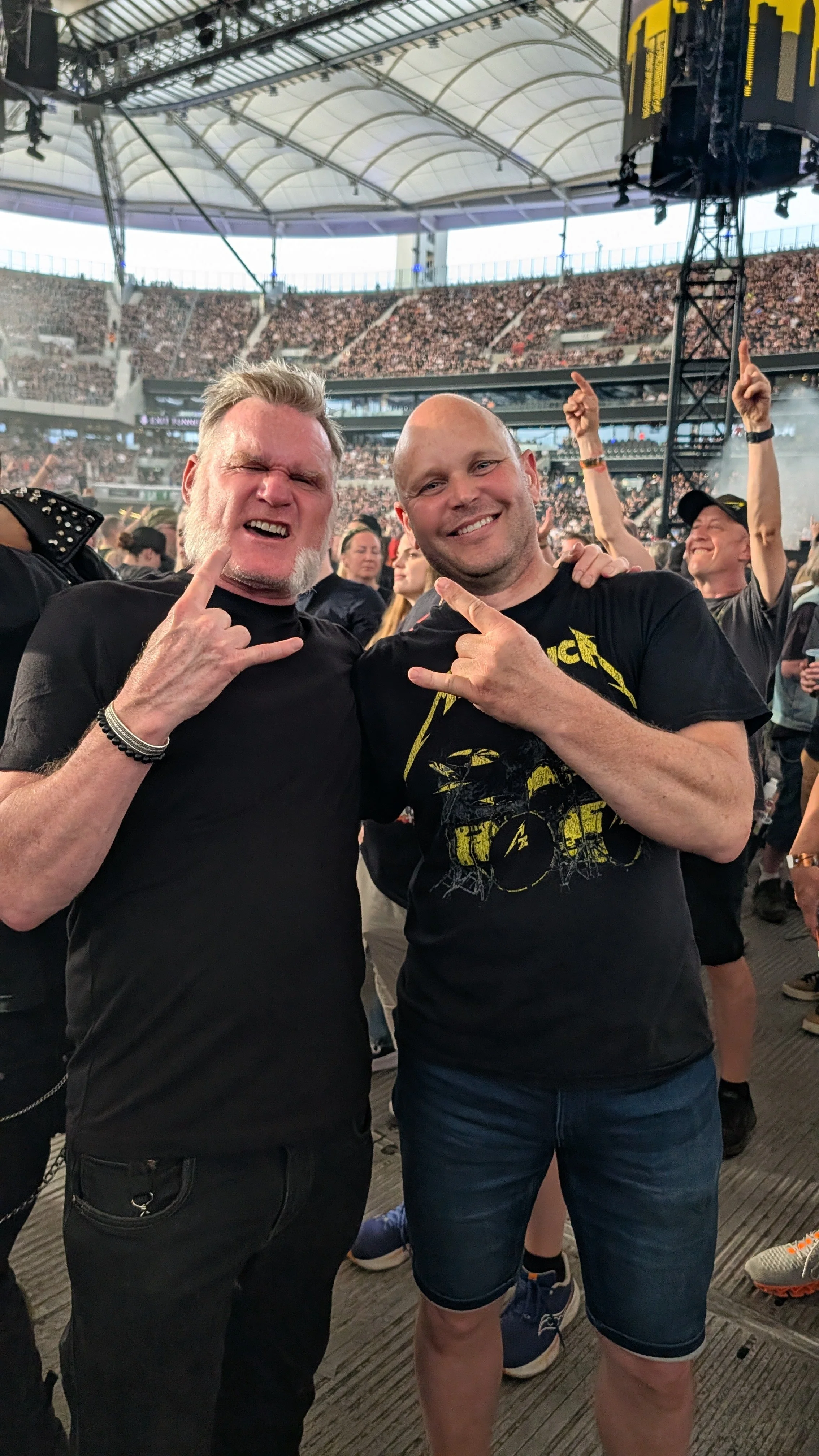 Two men at a concert or music festival, making rock and roll hand signs and smiling at the camera, with a large crowd and concert stage in the background.