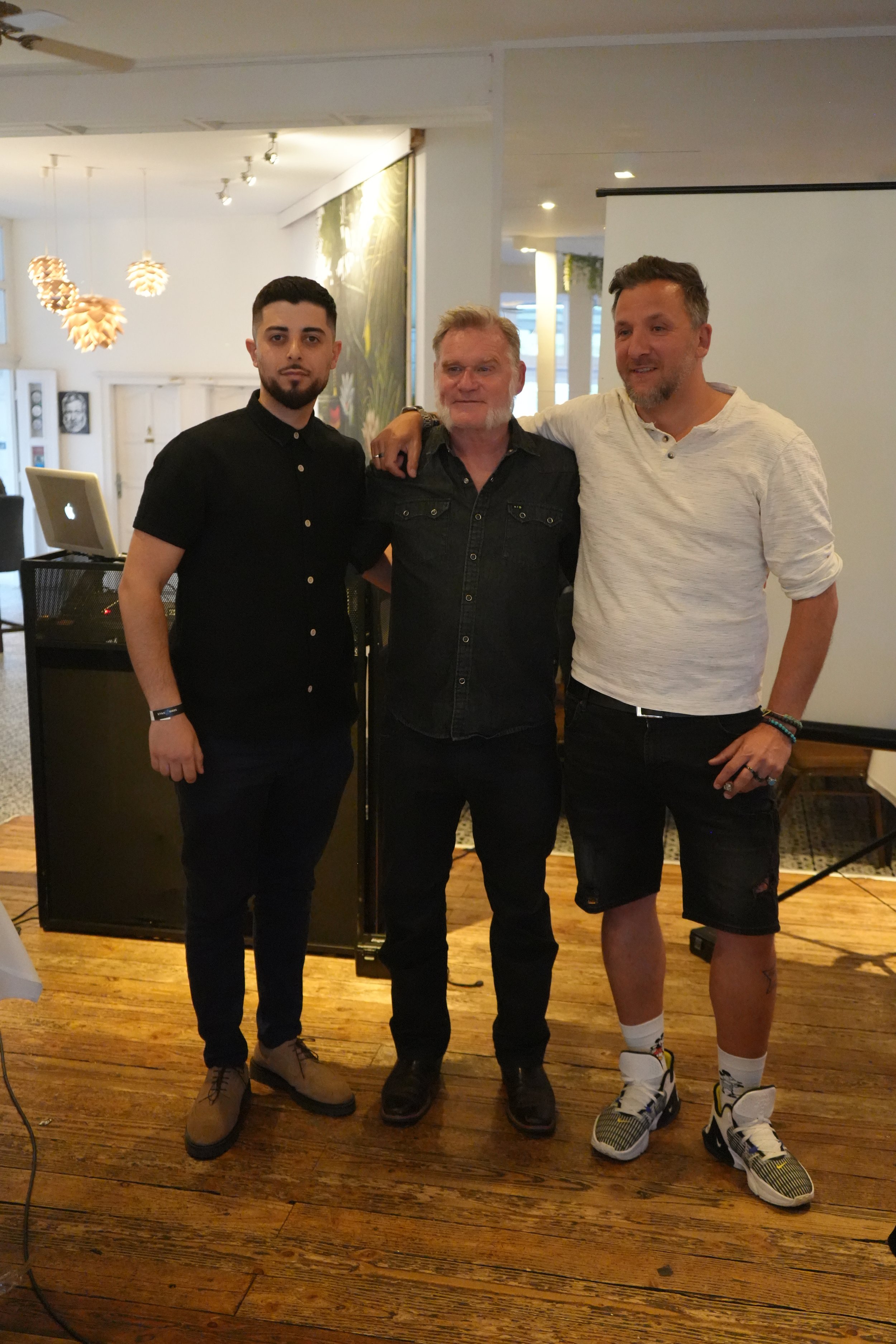 Three men standing side by side indoors, smiling for a photo. The man on the left is wearing a black shirt, the man in the middle is wearing a dark button-up shirt, and the man on the right is dressed in a white long-sleeve shirt and shorts.
